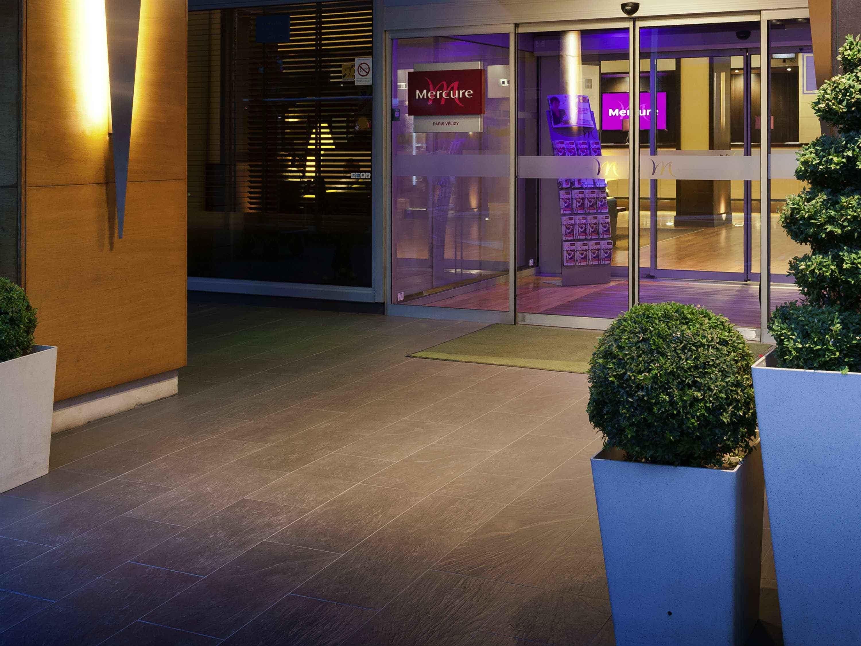 Una entrada moderna de edificio con puerta de vidrio, un cartel que dice 'Mercure Paris Velizy' y una cámara de seguridad encima de la puerta.