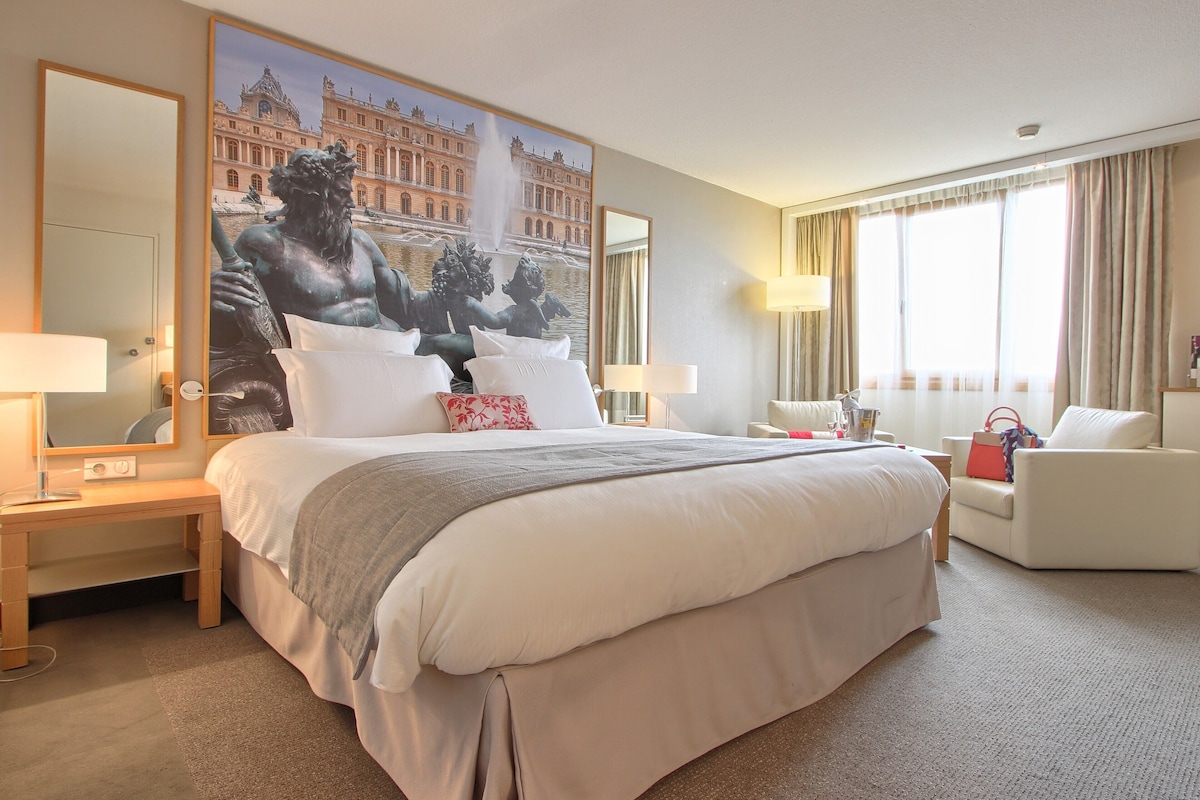 Une chambre d’hôtel avec un grand lit, un bureau, une chaise, une grande fenêtre avec des rideaux et un tableau représentant une fontaine.