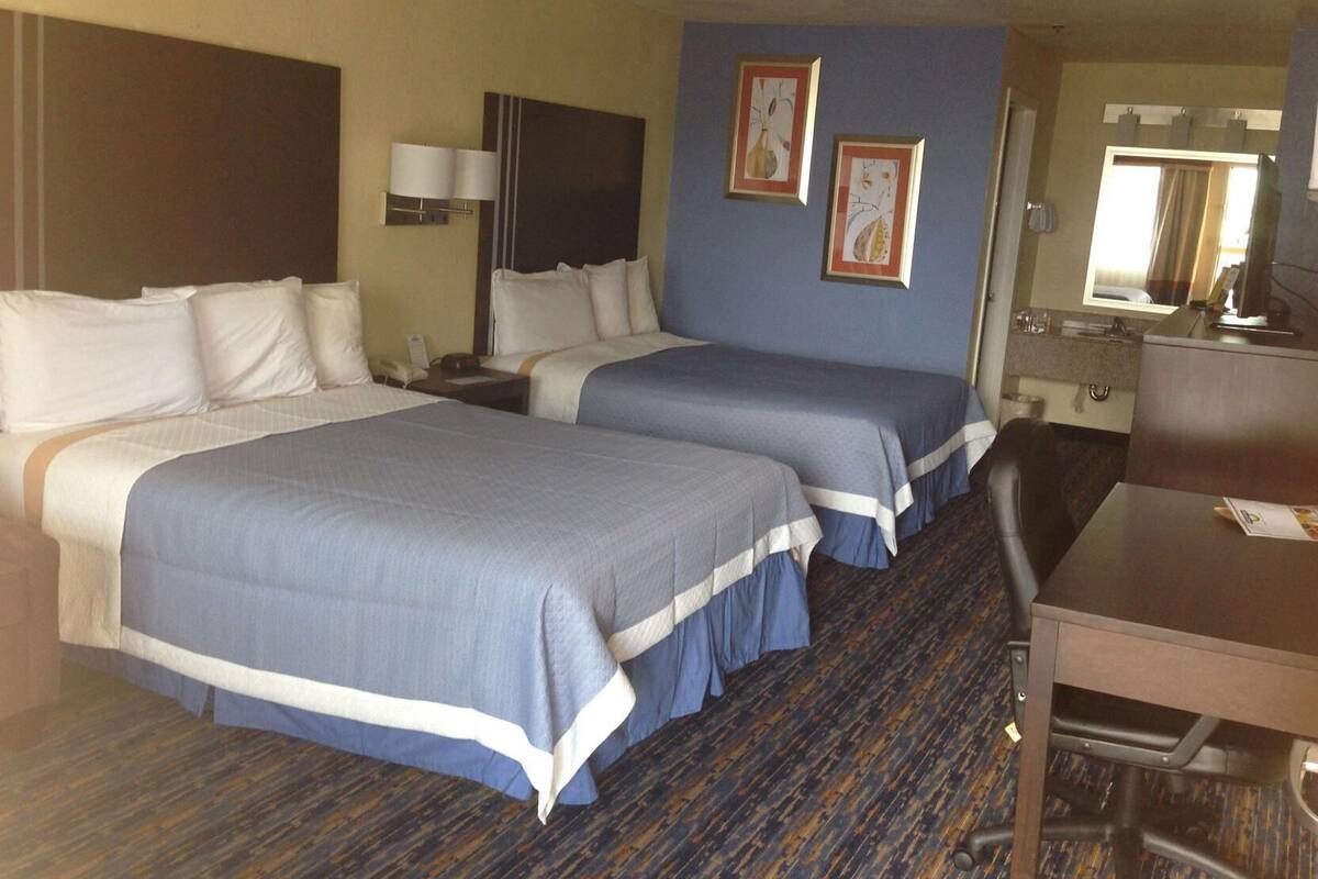 A hotel room with two beds, a desk, a chair, a nightstand, a lamp, a mirror, and two framed pictures on the wall.