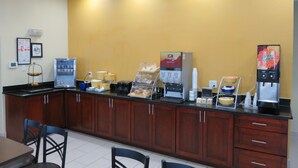 A breakfast bar with coffee machines, a water dispenser, and a fruit display in a room with wooden cabinets and a yellow wall. - Days Inn by Wyndham Near Seaworld San Antonio (San Antonio)
