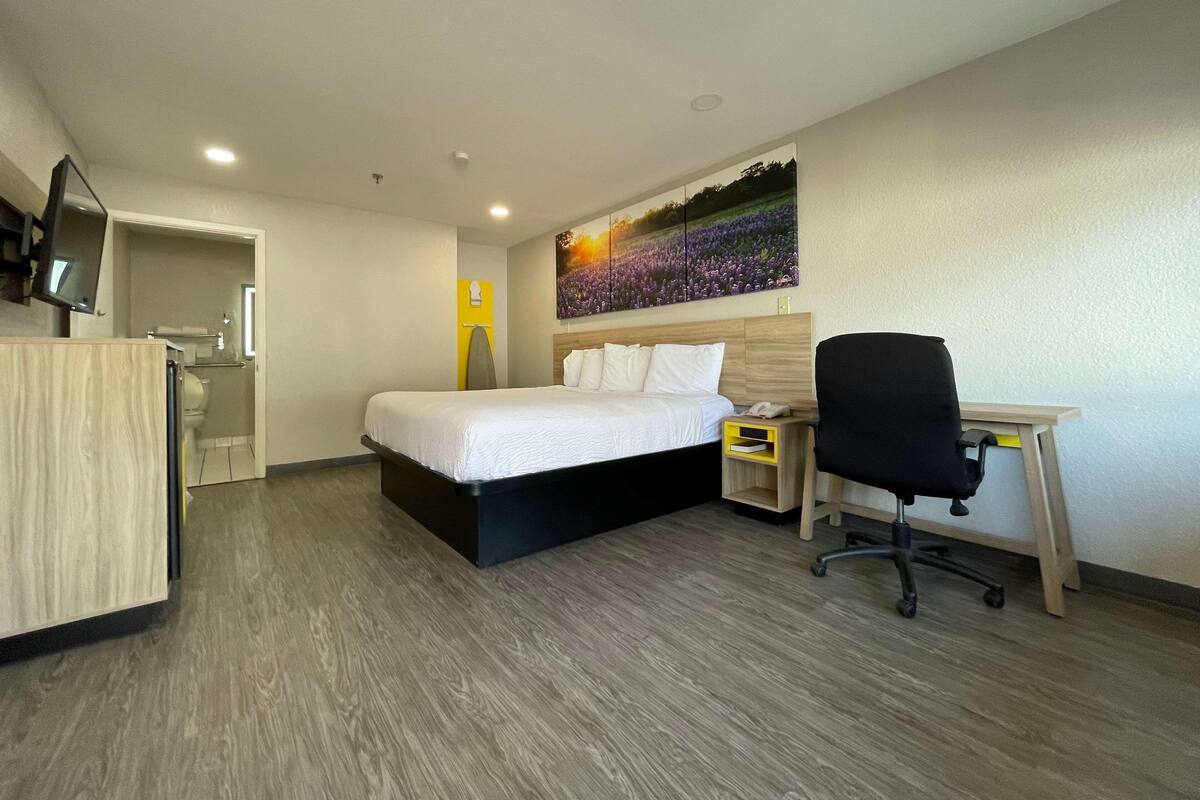 A hotel room with a bed, desk, chair, and a painting on the wall.