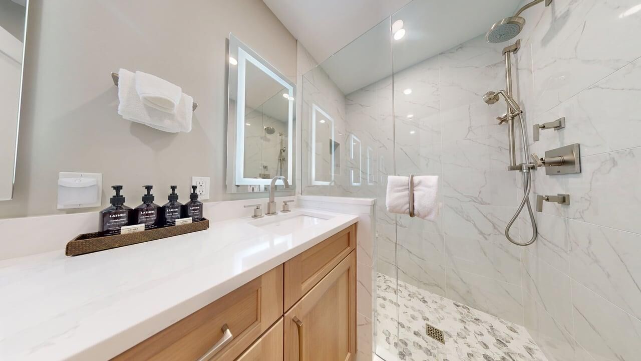 A modern bathroom with a white vanity, a large mirror, a shower with a handheld showerhead, and a marble-tiled floor.