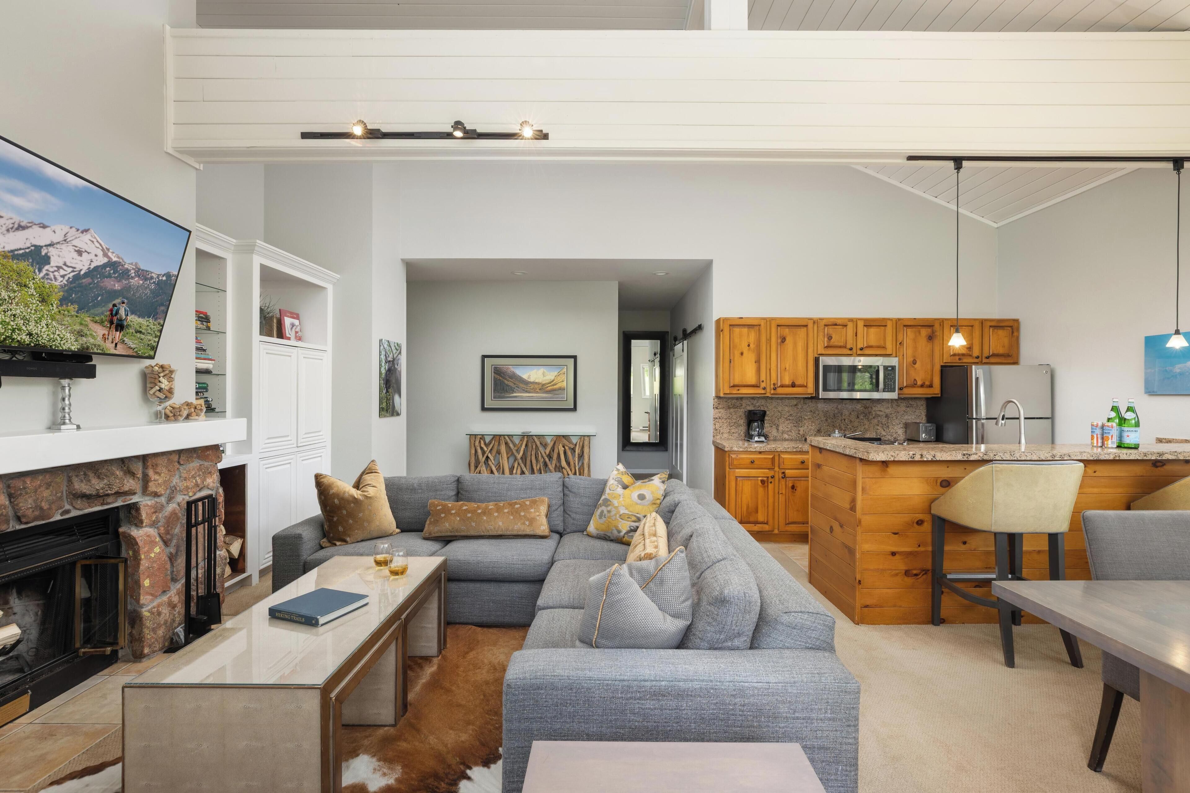 Uma sala de estar moderna com um sofá seccional cinza, uma lareira e uma área de cozinha com armários de madeira e bancada de granito.