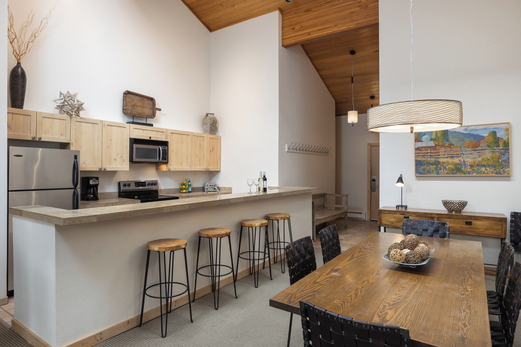 A modern kitchen with wooden cabinets, a stainless steel refrigerator, a microwave, and a wooden dining table with bar stools.