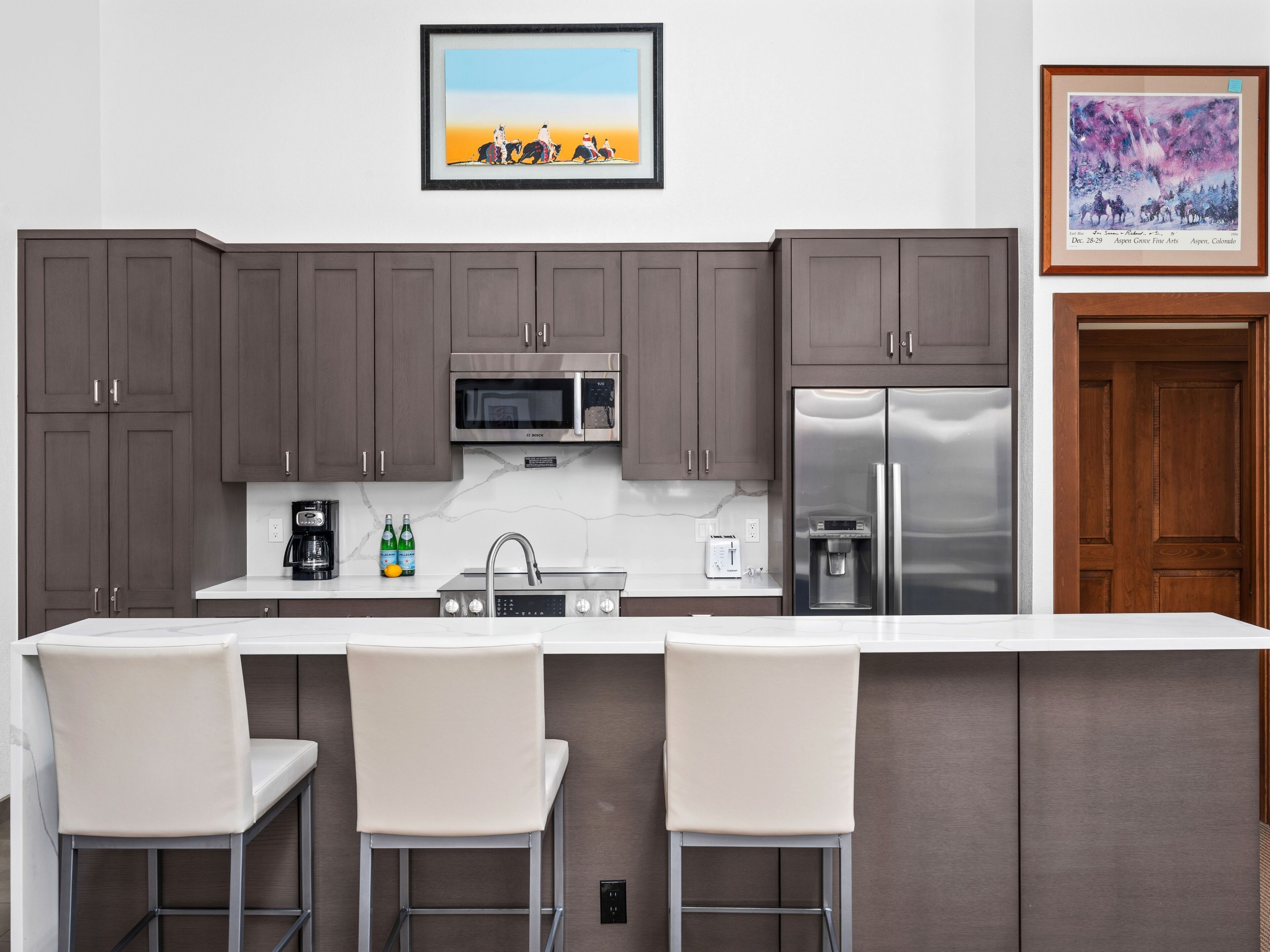 A modern kitchen with dark cabinets, stainless steel appliances, a marble countertop, and a bar area with two white chairs.