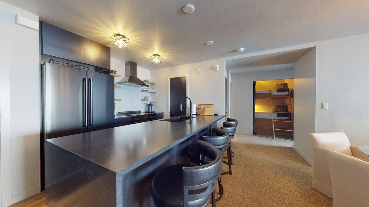 A modern kitchen with a large island, bar stools, and a built-in oven. There is a seating area with a white sofa and a small table.