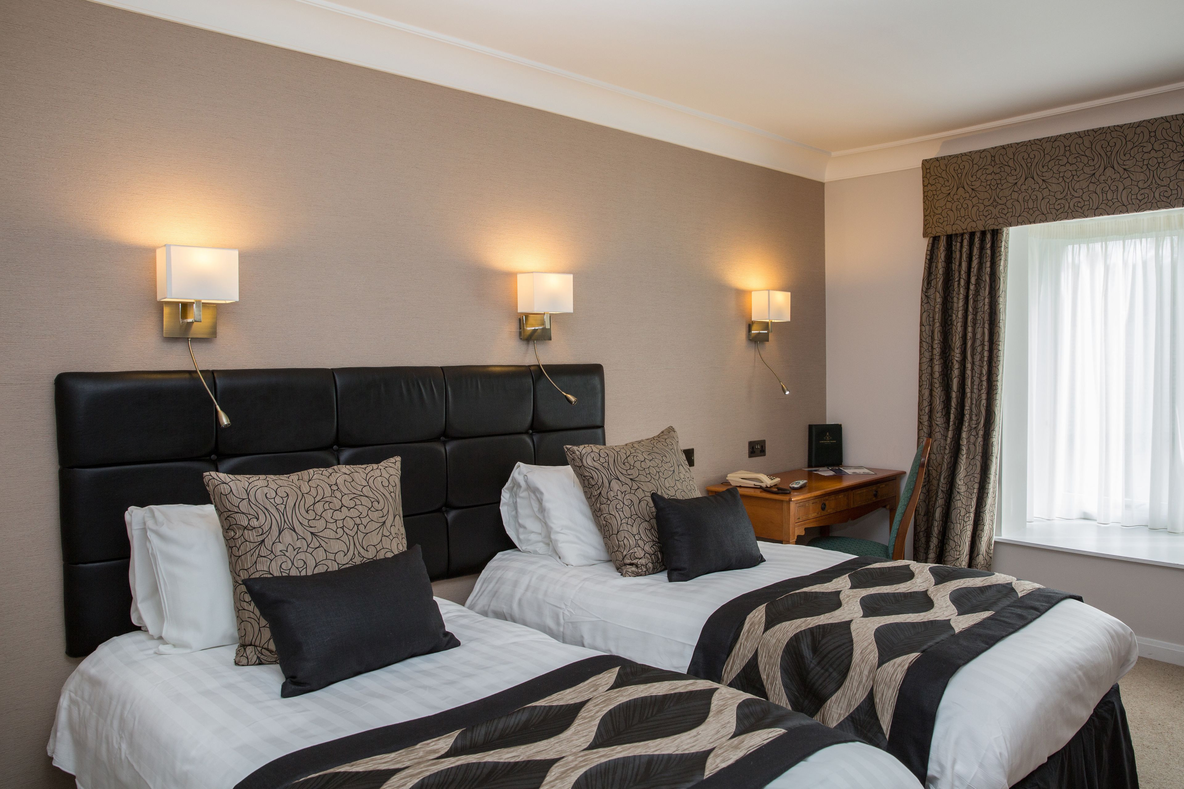 A hotel room with two beds, a headboard, bedside lamps, a desk, and a window with curtains.