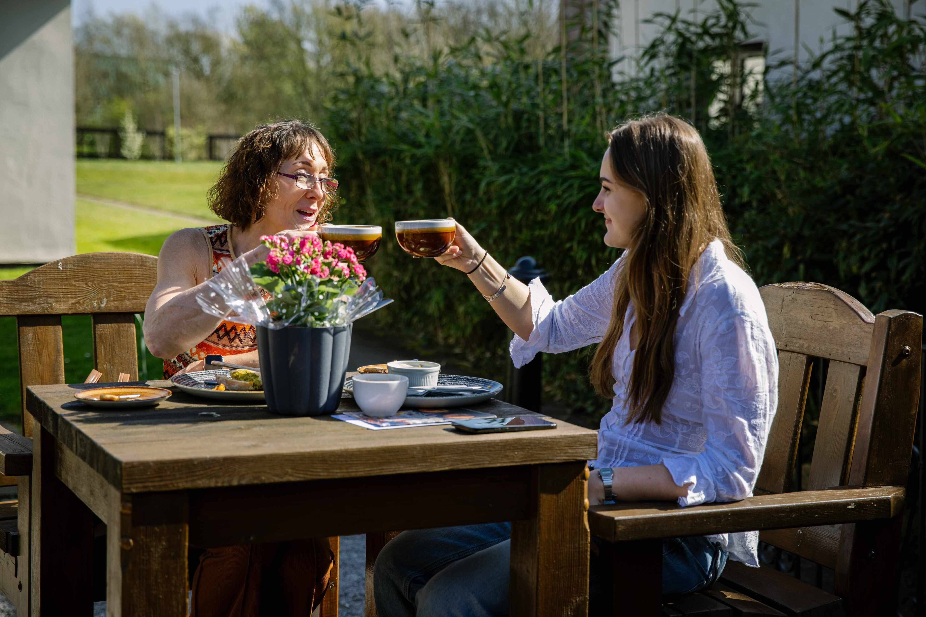 Deux personnes sont assises à une table en bois à l’extérieur, avec un jardin en arrière-plan.