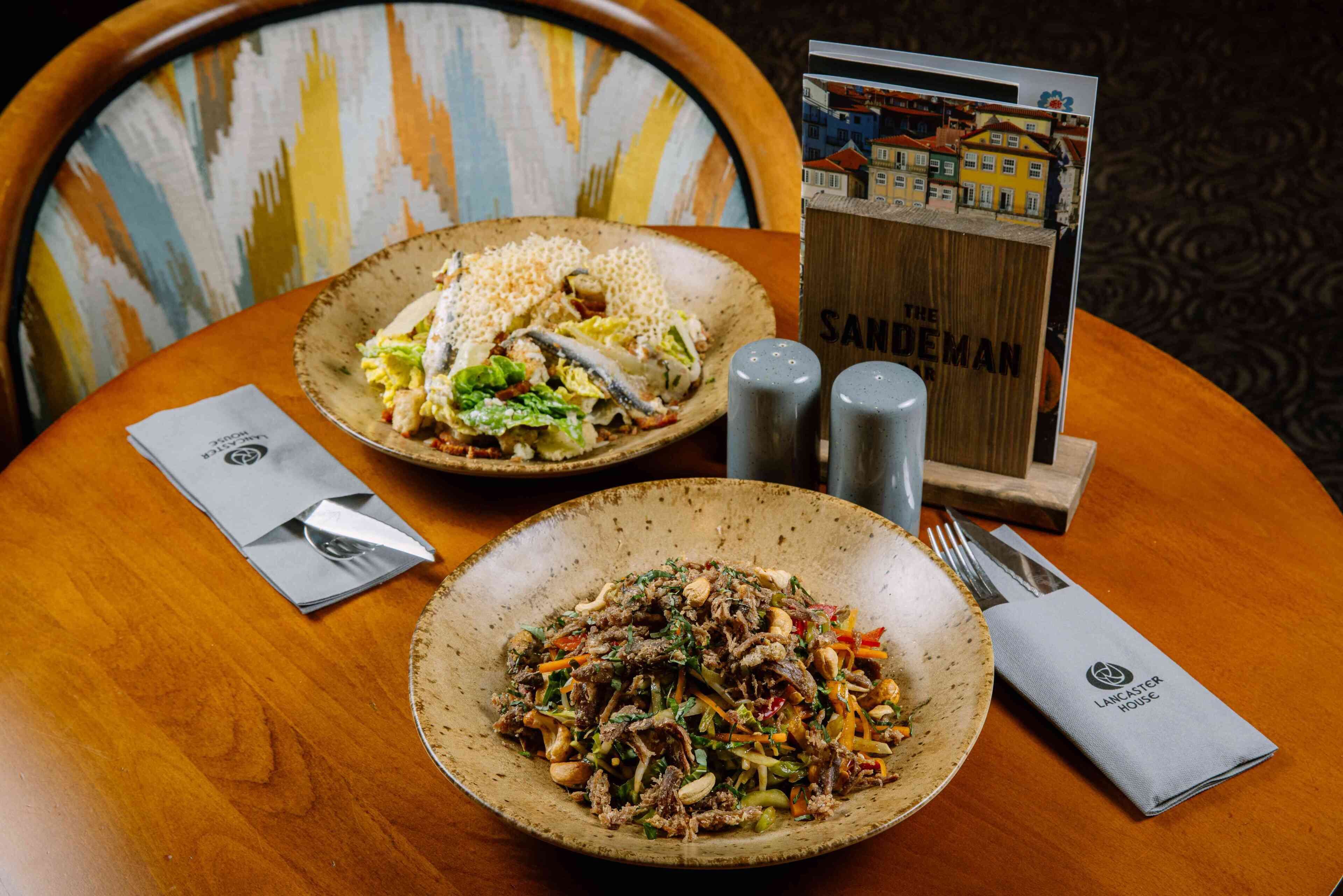 A dining table with two plates of food, salt and pepper shakers, a menu stand, and a napkin with a logo.