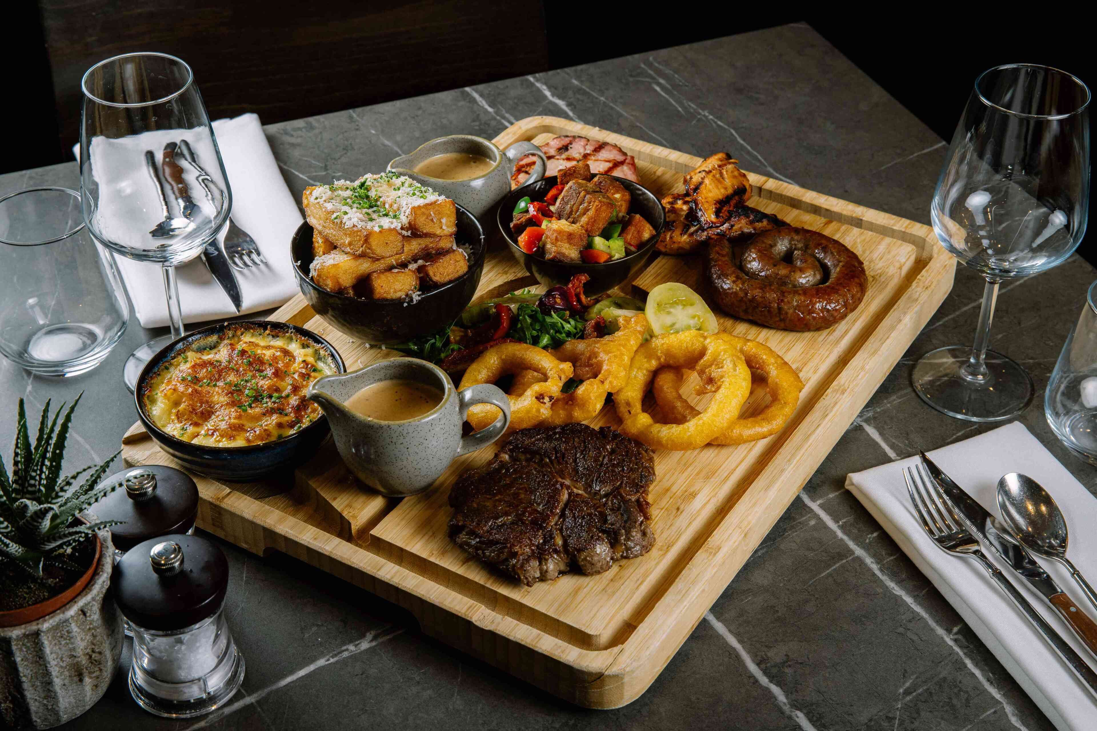 A wooden board with various dishes including a steak, onion rings, and a bowl of soup.
