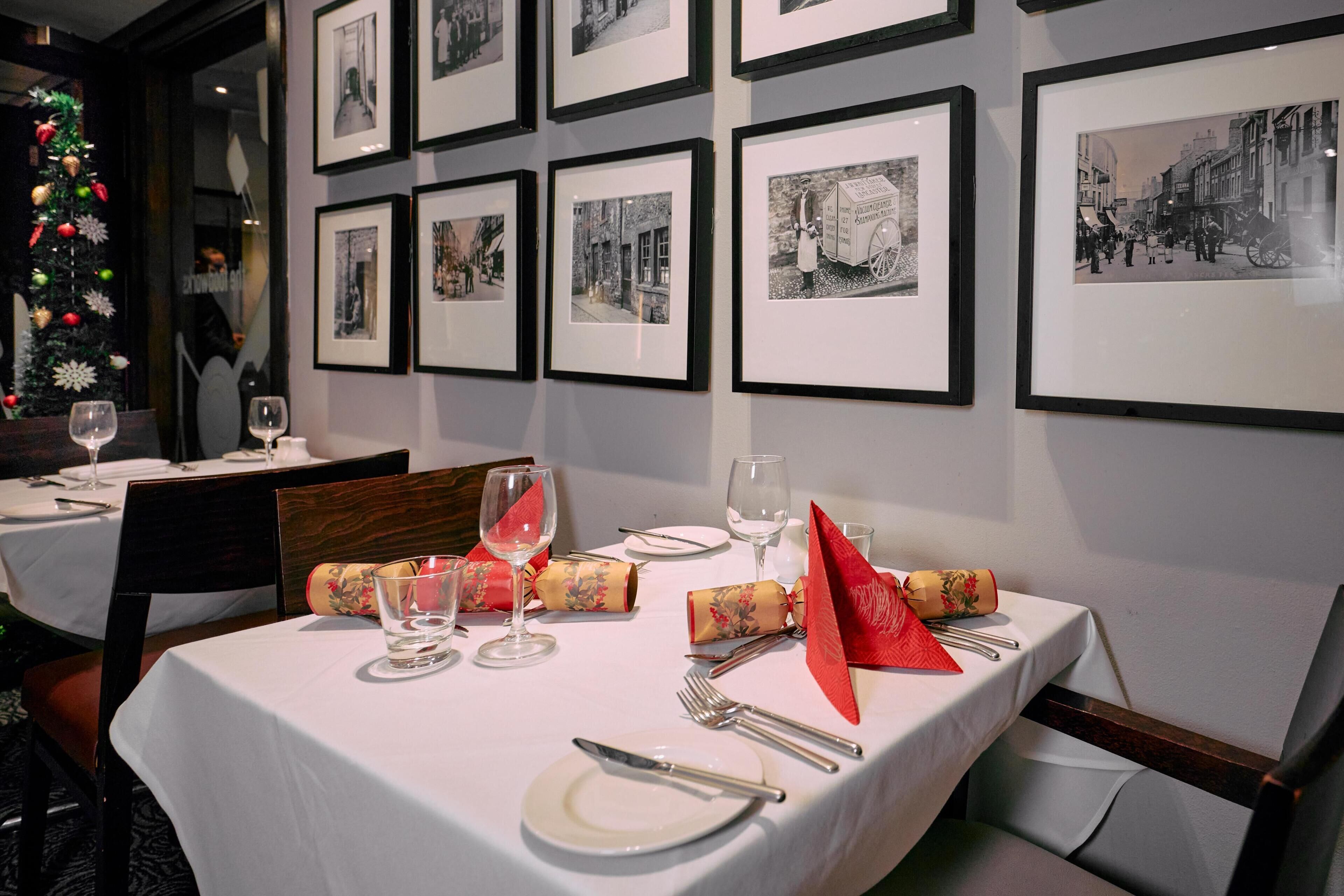 A dining table set with white tablecloth, plates, cutlery, and glasses, with framed black and white photographs on the wall.
