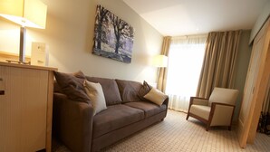 Quarto de hotel com sofá marrom, um armário de madeira, uma cadeira de madeira, um quadro na parede e um abajur.