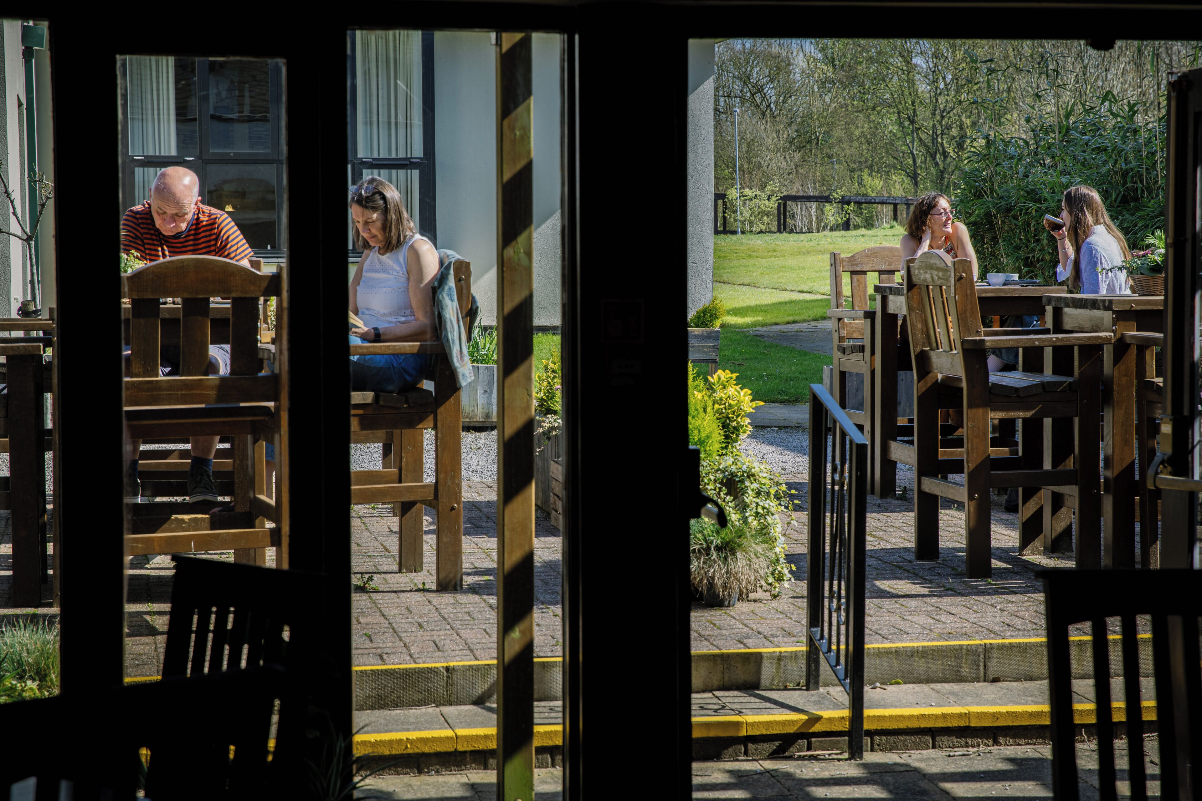 An outdoor seating area with wooden tables and chairs, a pathway leading to a grassy area, and a building entrance.
