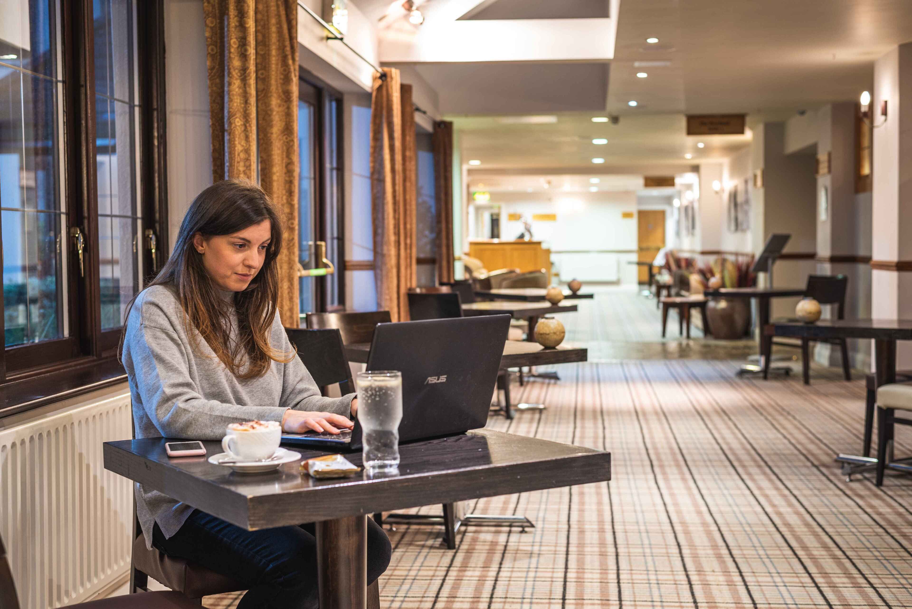 Une personne est assise à une table dans le hall d’un hôtel, avec un ordinateur portable, un verre d’eau et une assiette de nourriture devant elle. On aperçoit en arrière-plan le comptoir d’enregistrement.