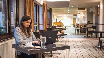Una persona sentada a una mesa con una computadora portátil, un vaso de agua y un plato de comida en el vestíbulo de un hotel, con un mostrador de recepción al fondo.