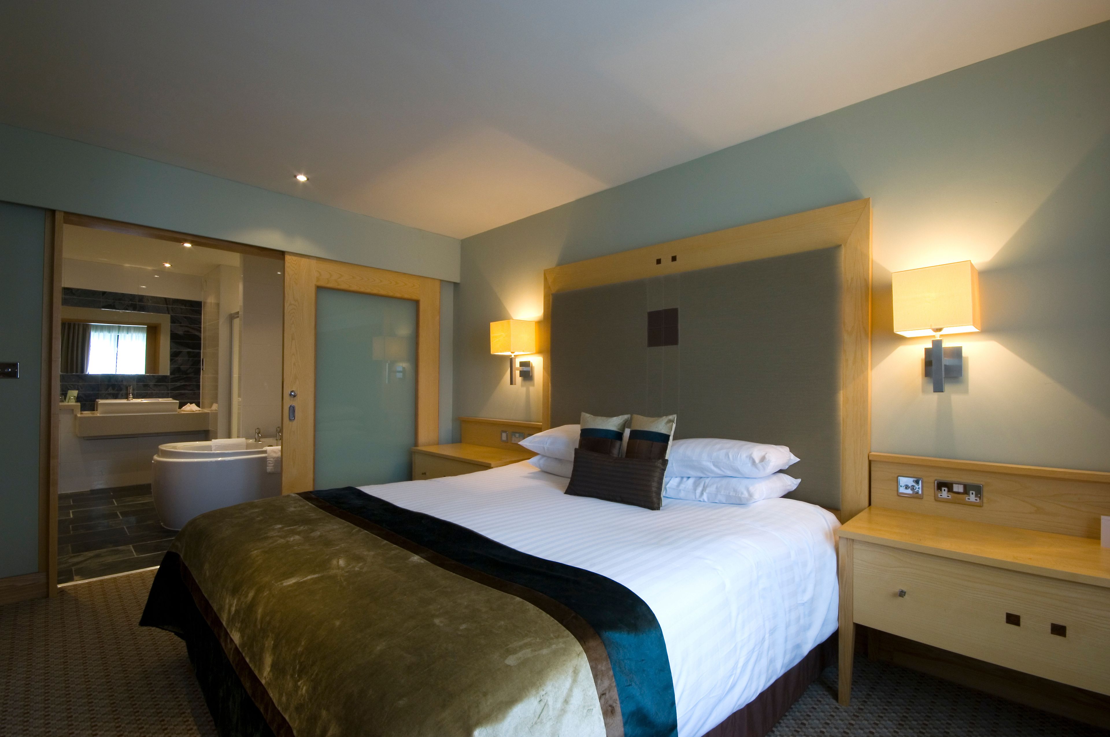 A hotel room with a large bed, a wooden nightstand, a bedside table, a wall-mounted lamp, and a bathroom visible through an open door.