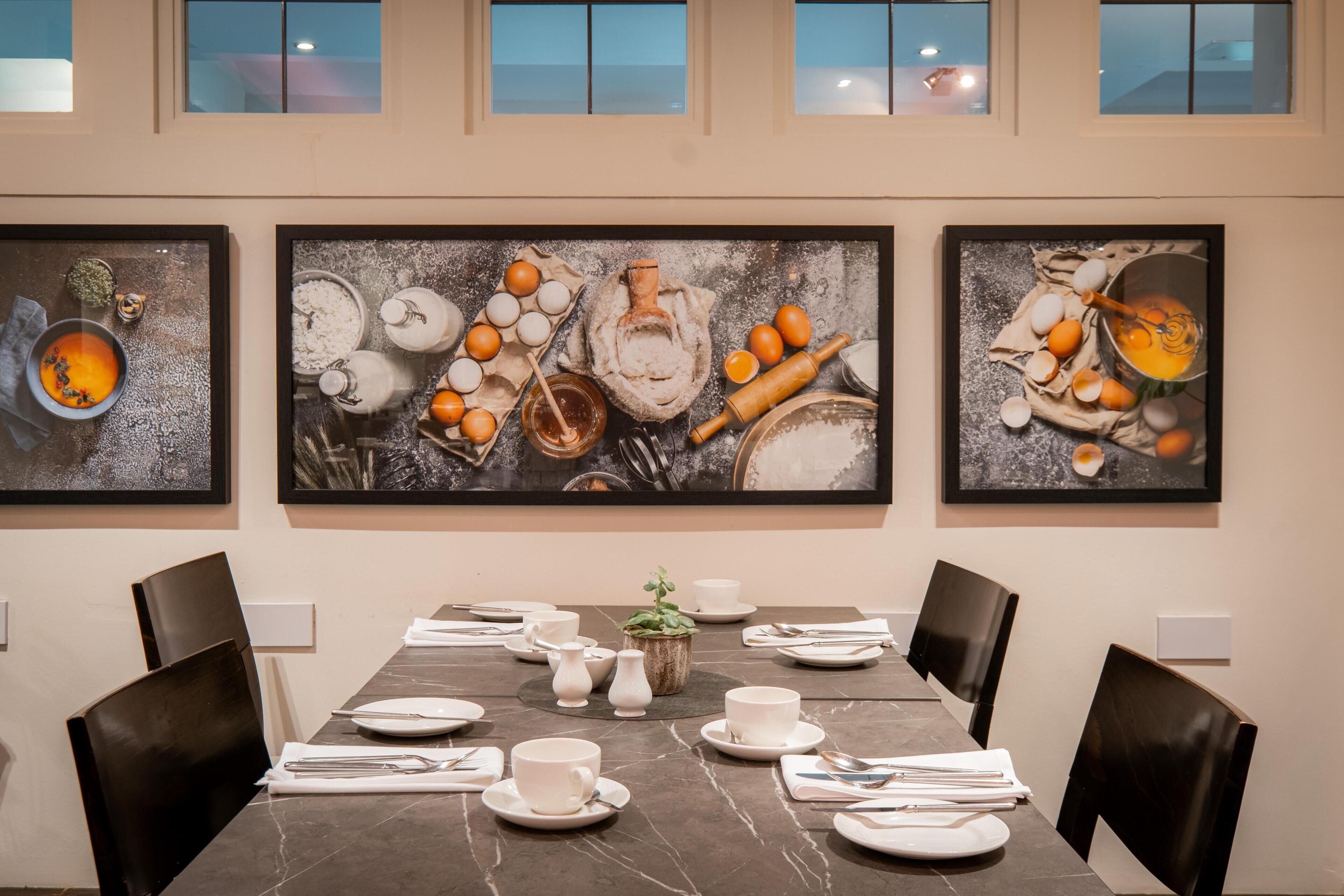 A dining area with a marble table, white plates, cups, and cutlery. There are two framed pictures on the wall depicting food items.