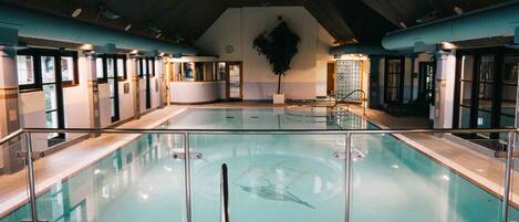 Una amplia piscina interior con paredes de vidrio transparente, suelo de baldosas y techo con vigas a la vista.