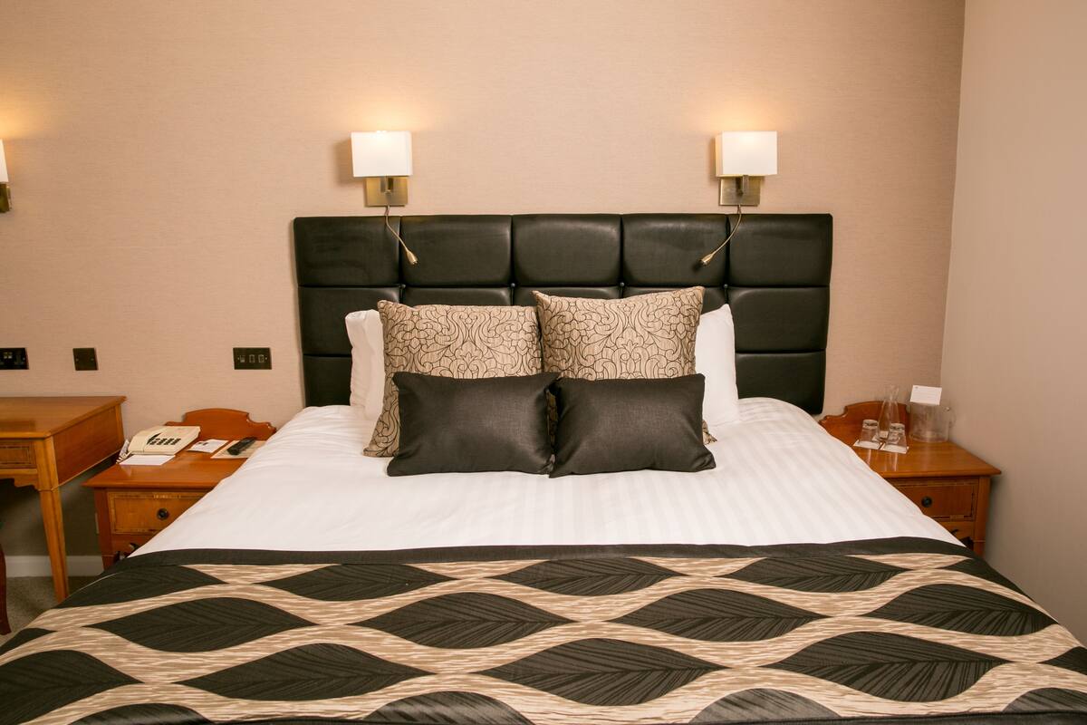 A neatly made bed with a black and beige patterned bedspread, two matching pillows, and a black upholstered headboard. There are two bedside tables with lamps, a wooden nightstand, and a telephone.