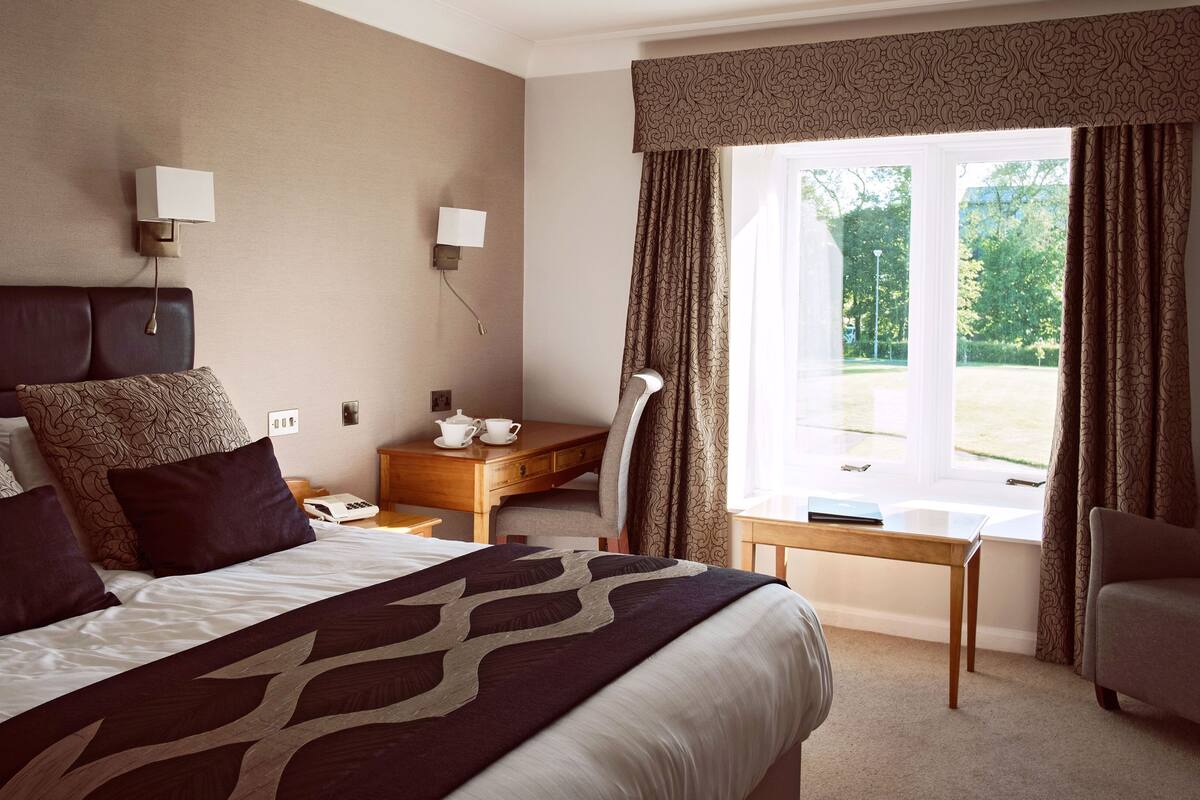 A hotel room with a large bed, a desk with a chair, a window with curtains, and a view of the outdoors.