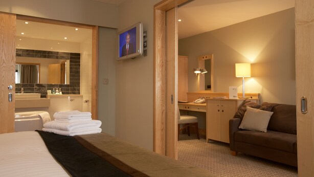 Quarto de hotel com cama, escrivaninha, cadeira, sofá, televisão e banheiro com espelho e pia.