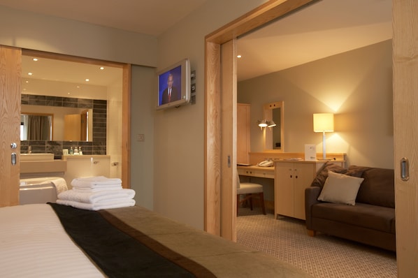 A hotel room with a bed, a desk, a chair, a sofa, a TV, and a bathroom with a mirror and sink.