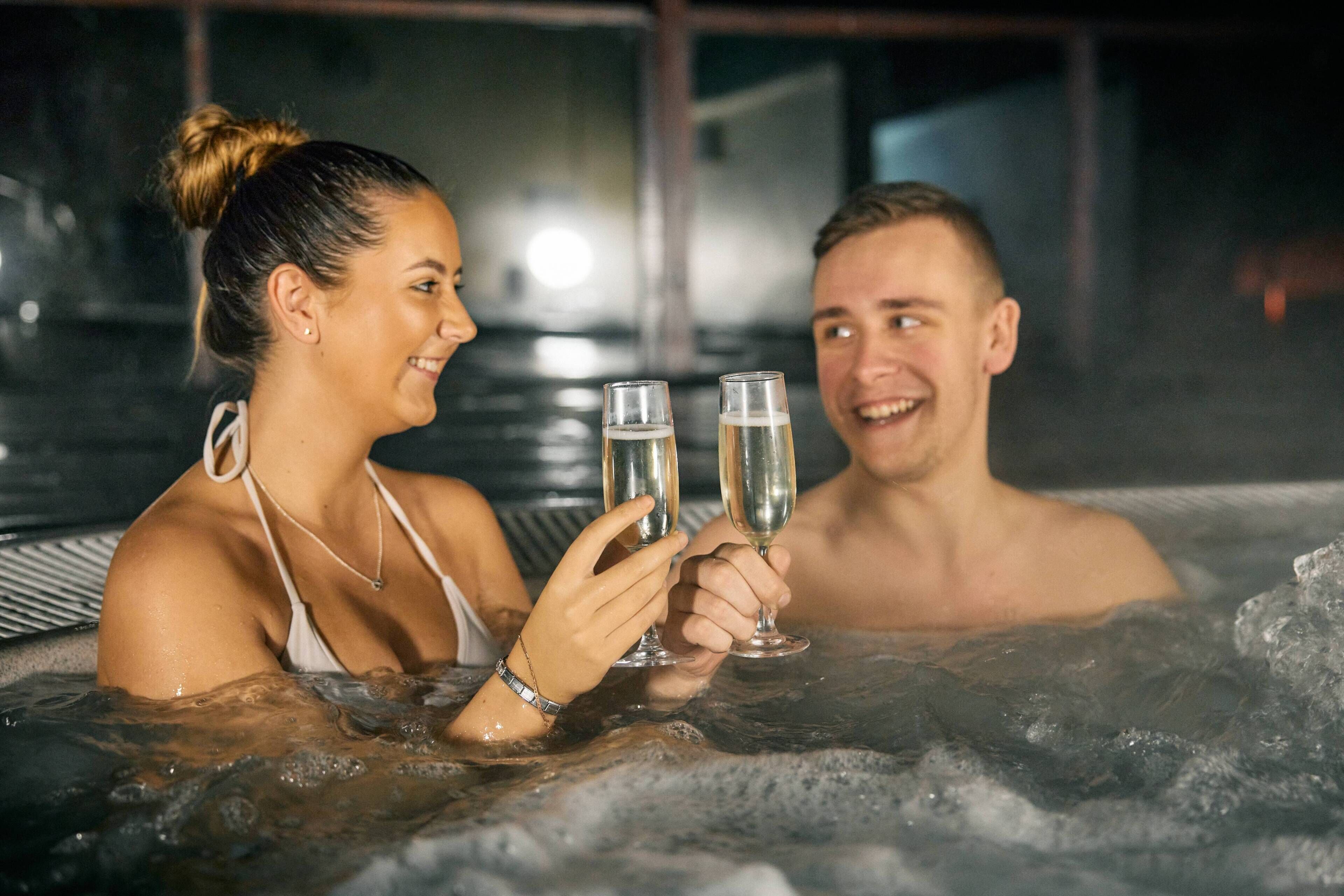 Dos personas en un jacuzzi sosteniendo vasos, probablemente con bebida.