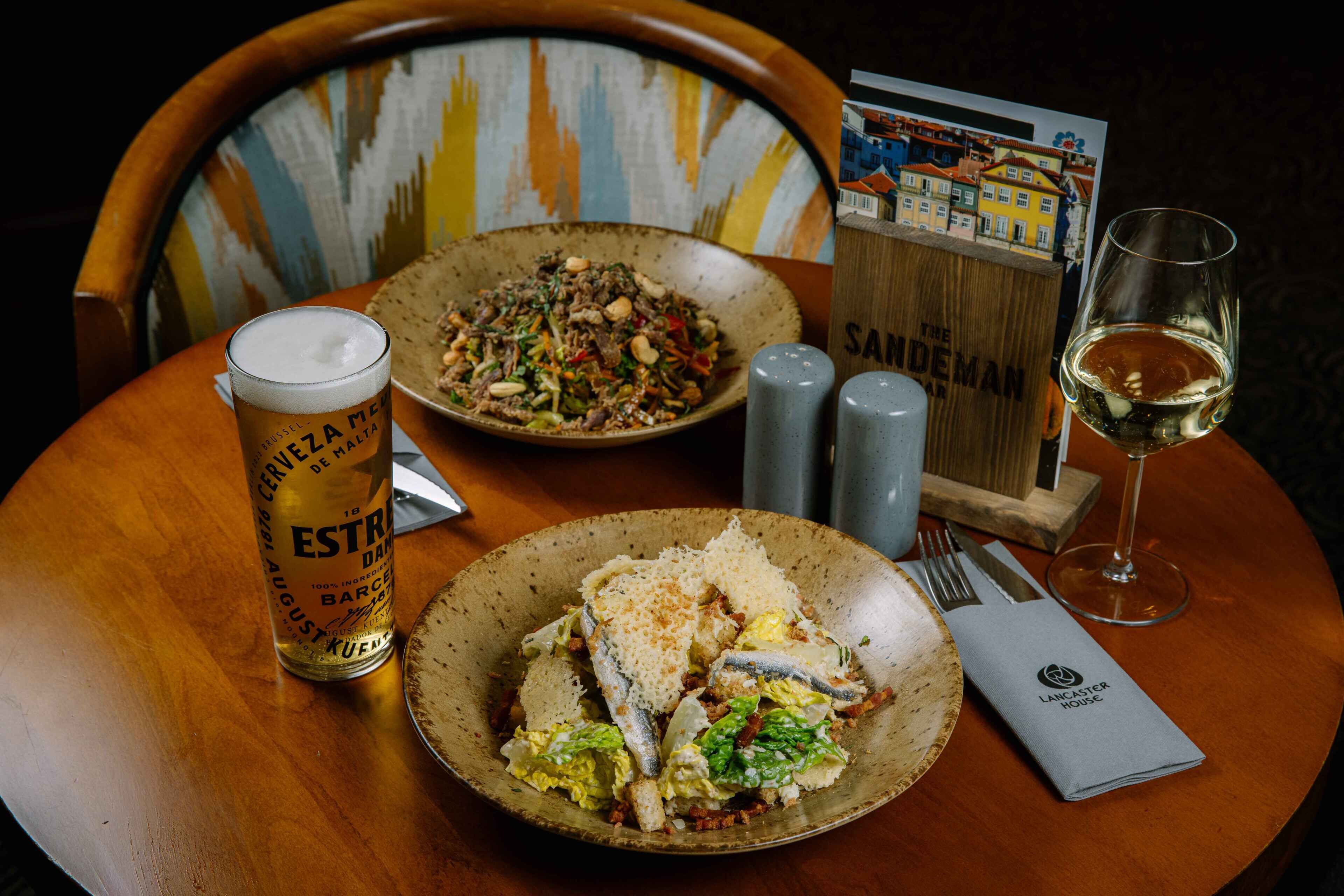 A dining table with a beer glass, a plate of fish and salad, a wine glass, salt and pepper shakers, and a menu.