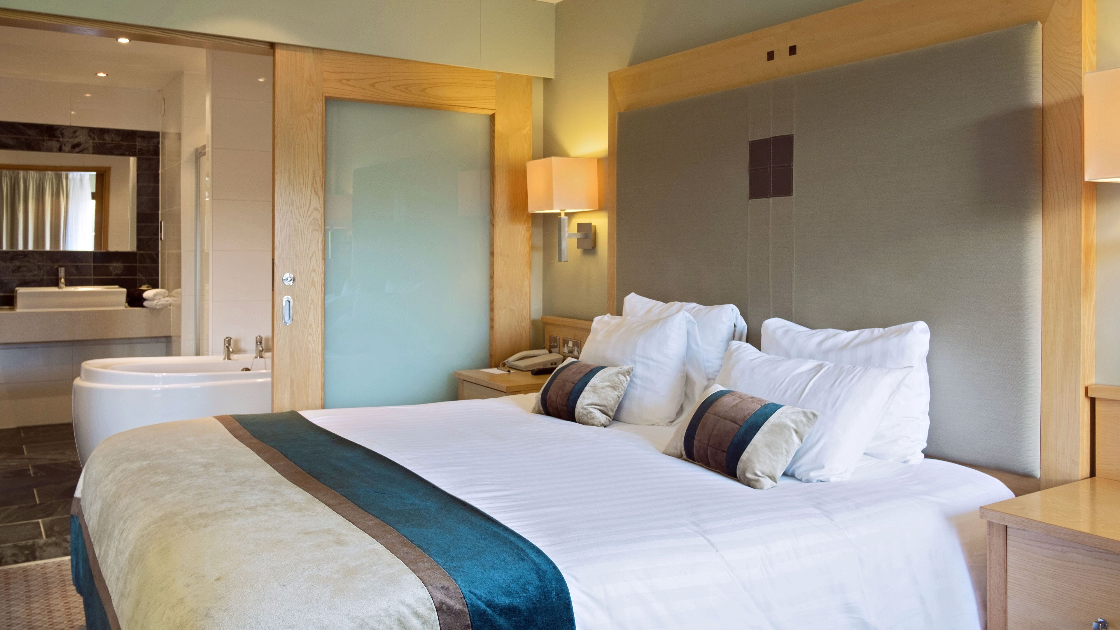 Habitación de hotel con una cama grande, una mesita de noche, una lámpara, un espejo y un baño con lavamanos y bañera.