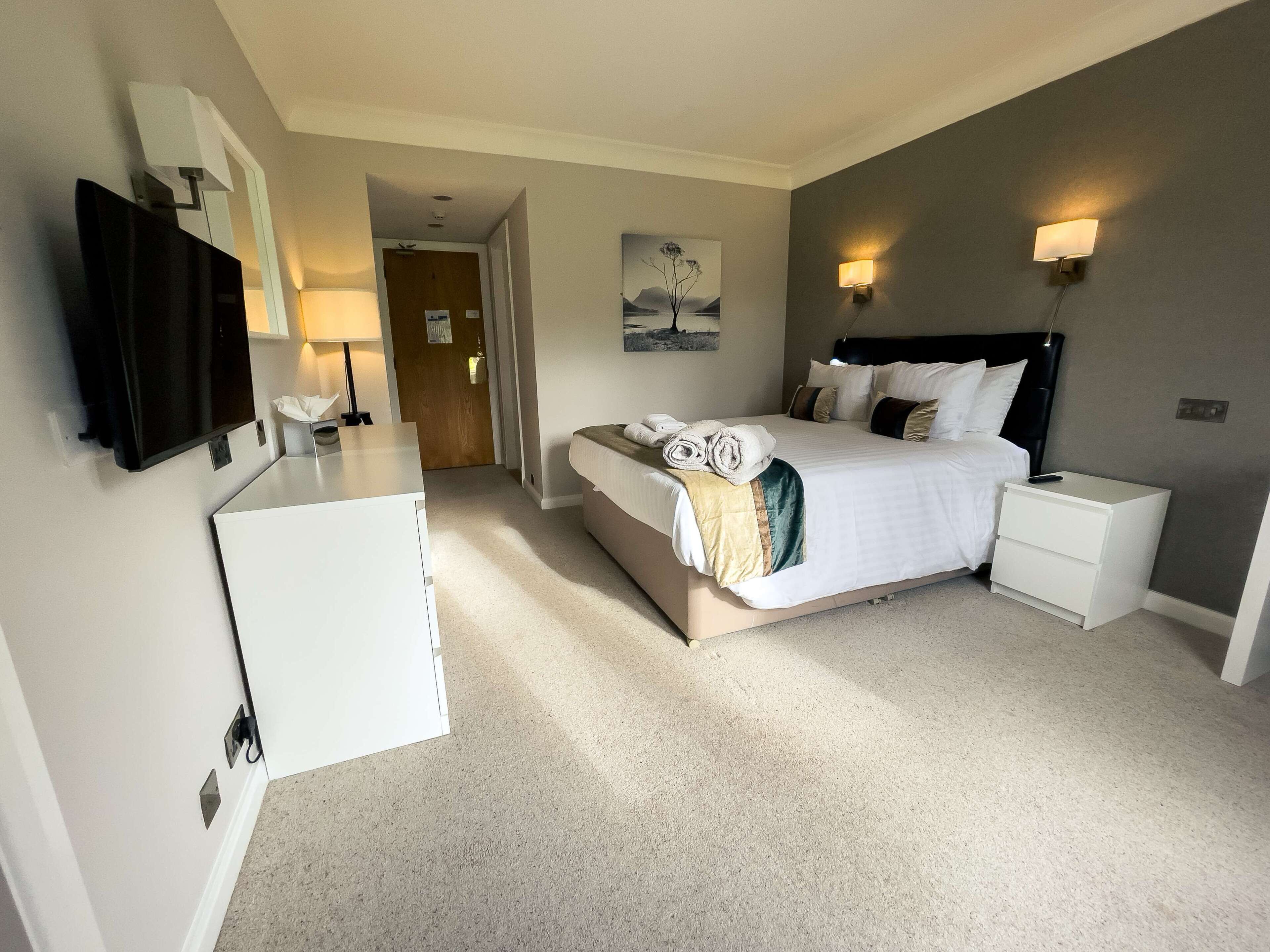 A hotel room with a bed, bedside tables, a TV, and a painting on the wall.