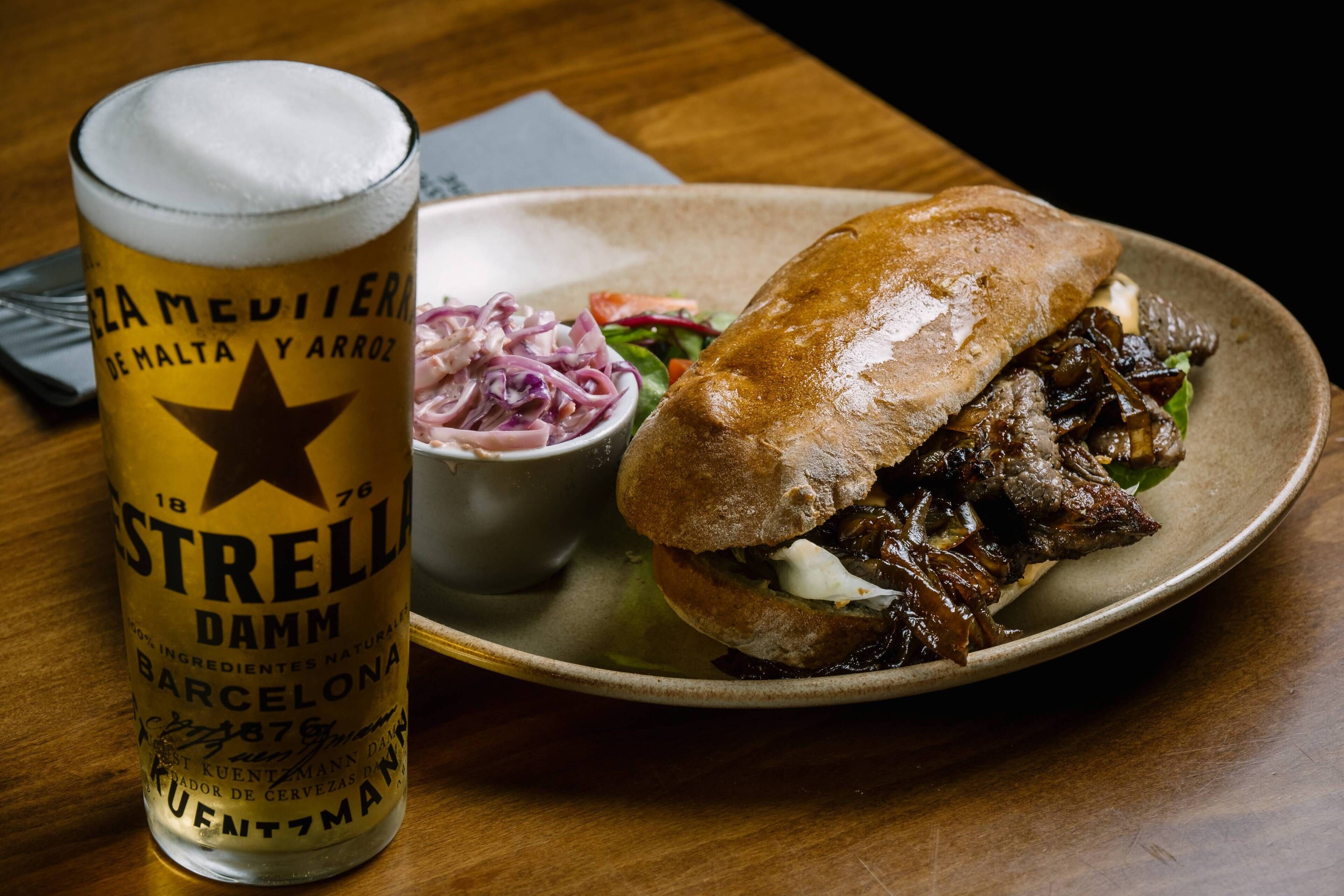A meal consisting of a sandwich with meat and vegetables, a side of red onion salad, and a glass of beer.