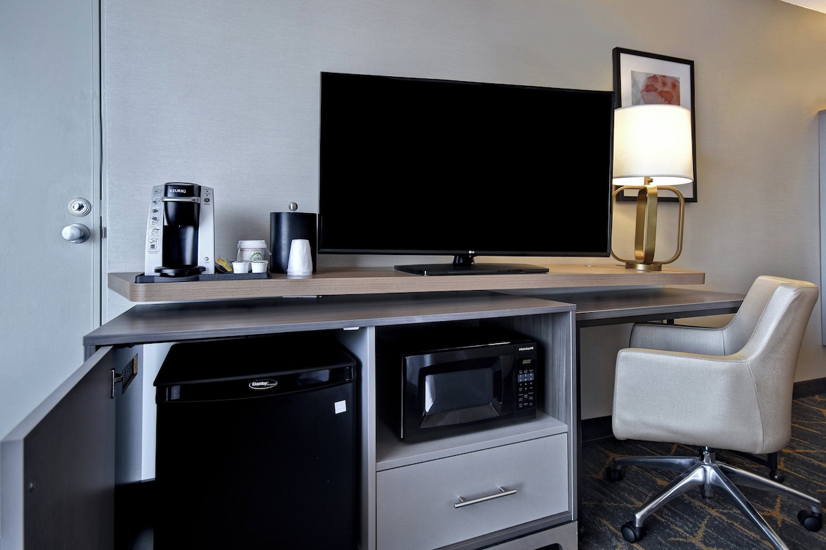 Quarto de hotel com escrivaninha, televisão de tela plana, micro-ondas, cafeteira, luminária e cadeira.
