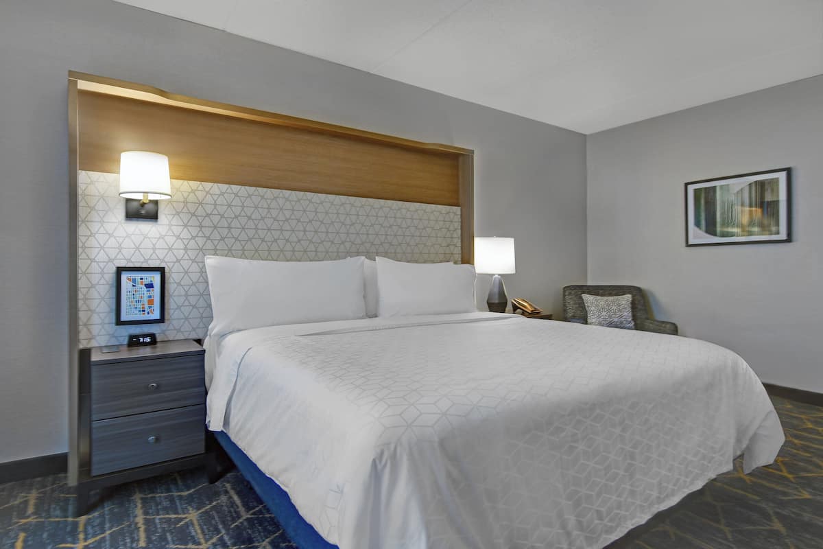 A hotel room with a large bed, a nightstand with a lamp, a chair, and a framed picture on the wall.