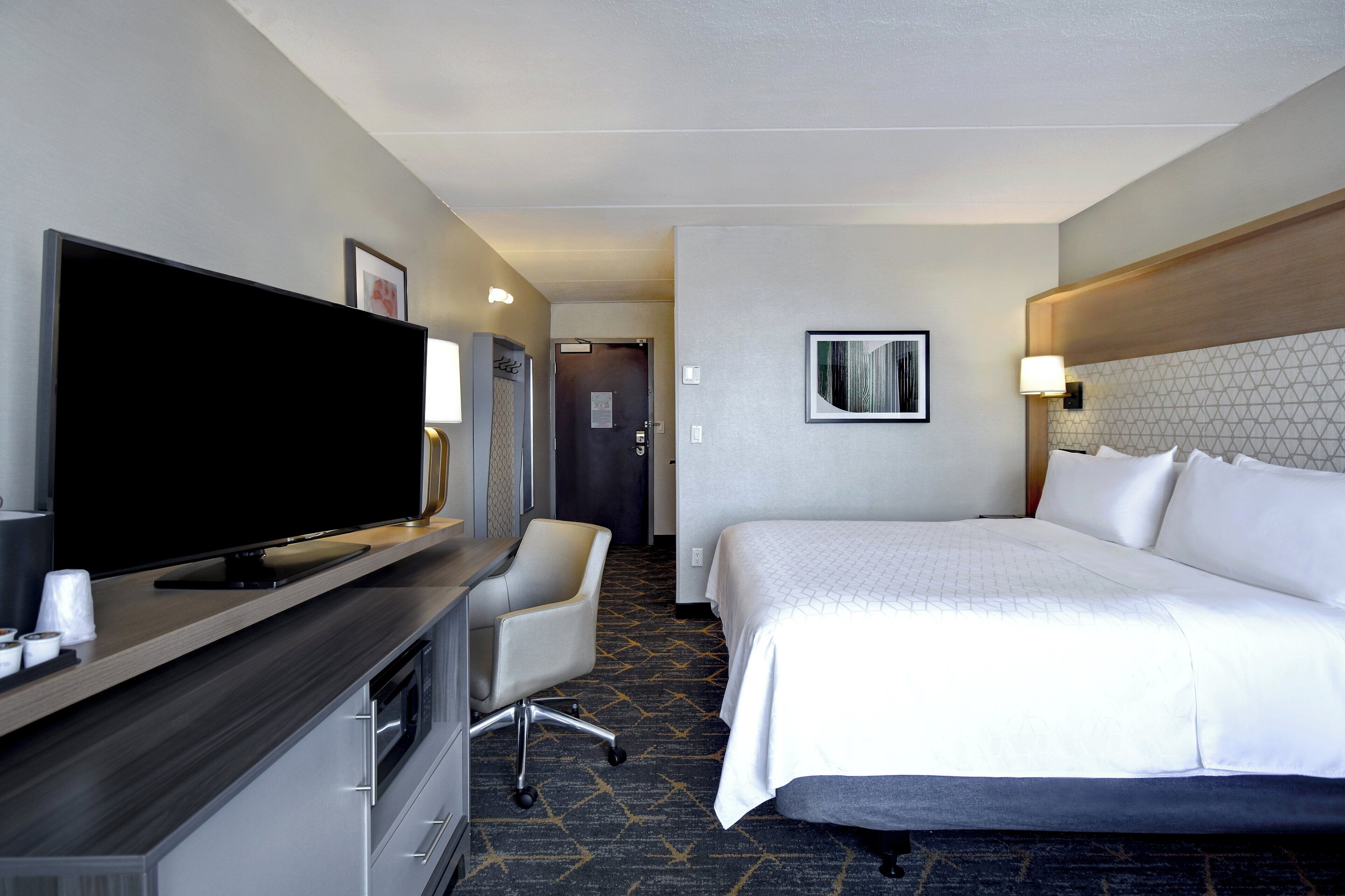 A hotel room with a bed, a desk with a flat-screen TV, a chair, and a wall-mounted lamp.