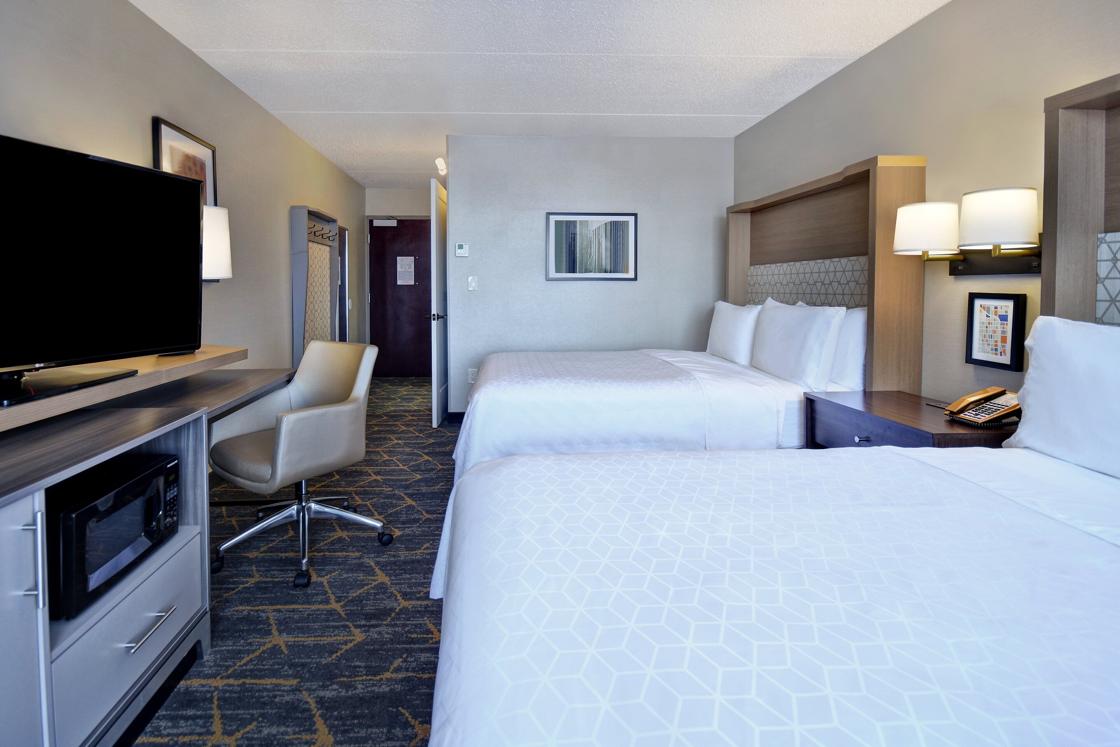A hotel room with a flat-screen TV, a desk with a microwave, two beds, a chair, and a door leading to another room.