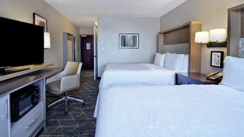 A hotel room with a flat-screen TV, a desk with a microwave, two beds, a chair, and a door leading to another room.
