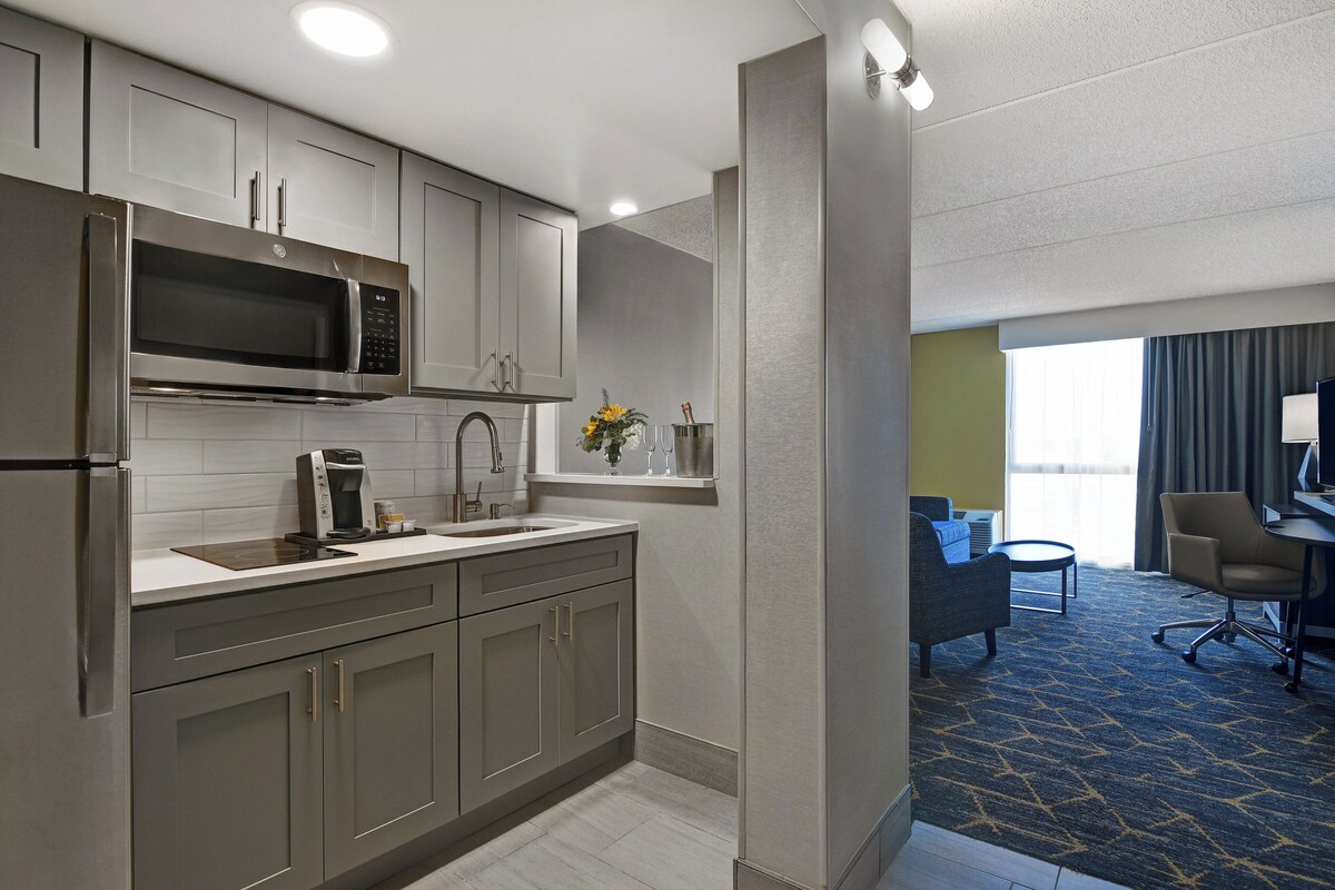 A modern kitchen with stainless steel appliances, a coffee maker, and a vase with flowers. There is a dining area with a sofa and a chair.