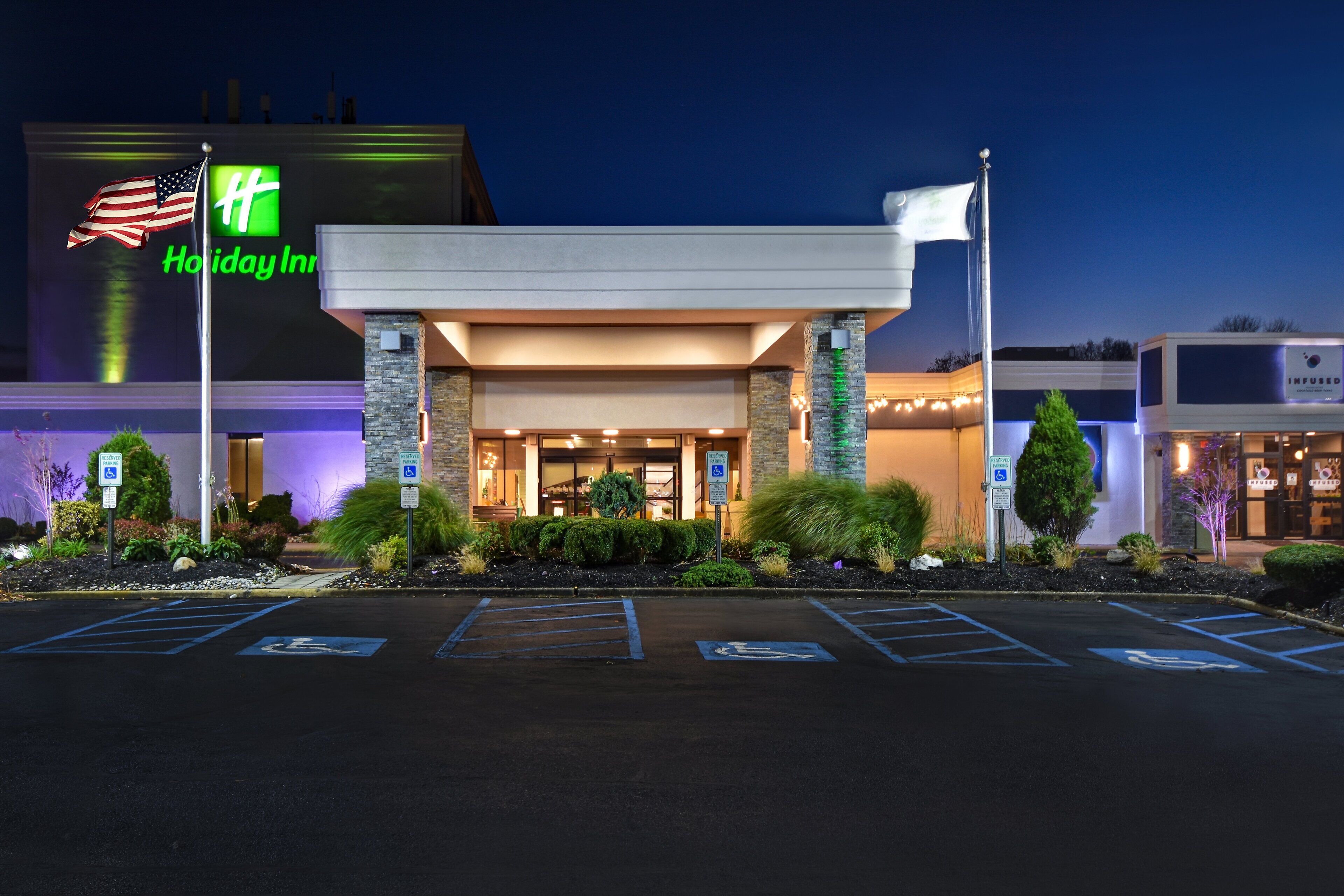 A Holiday Inn hotel entrance with a large sign, parking spaces, and a flagpole with an American flag.