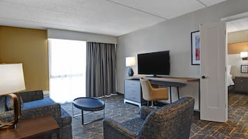 A hotel room with a desk, two chairs, a TV, a lamp, and a bed.