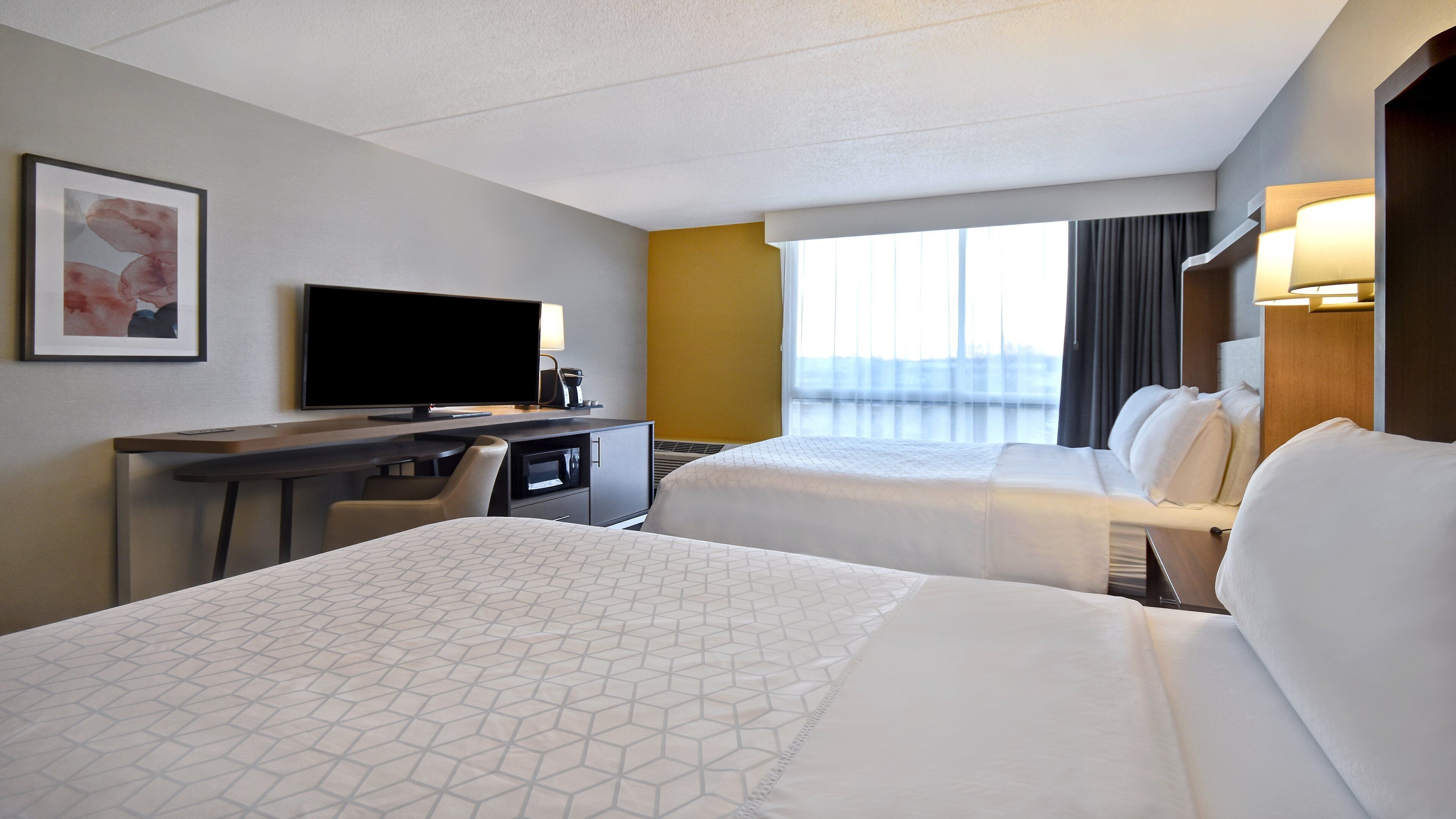 A hotel room with two beds, a desk with a television, and a large window.