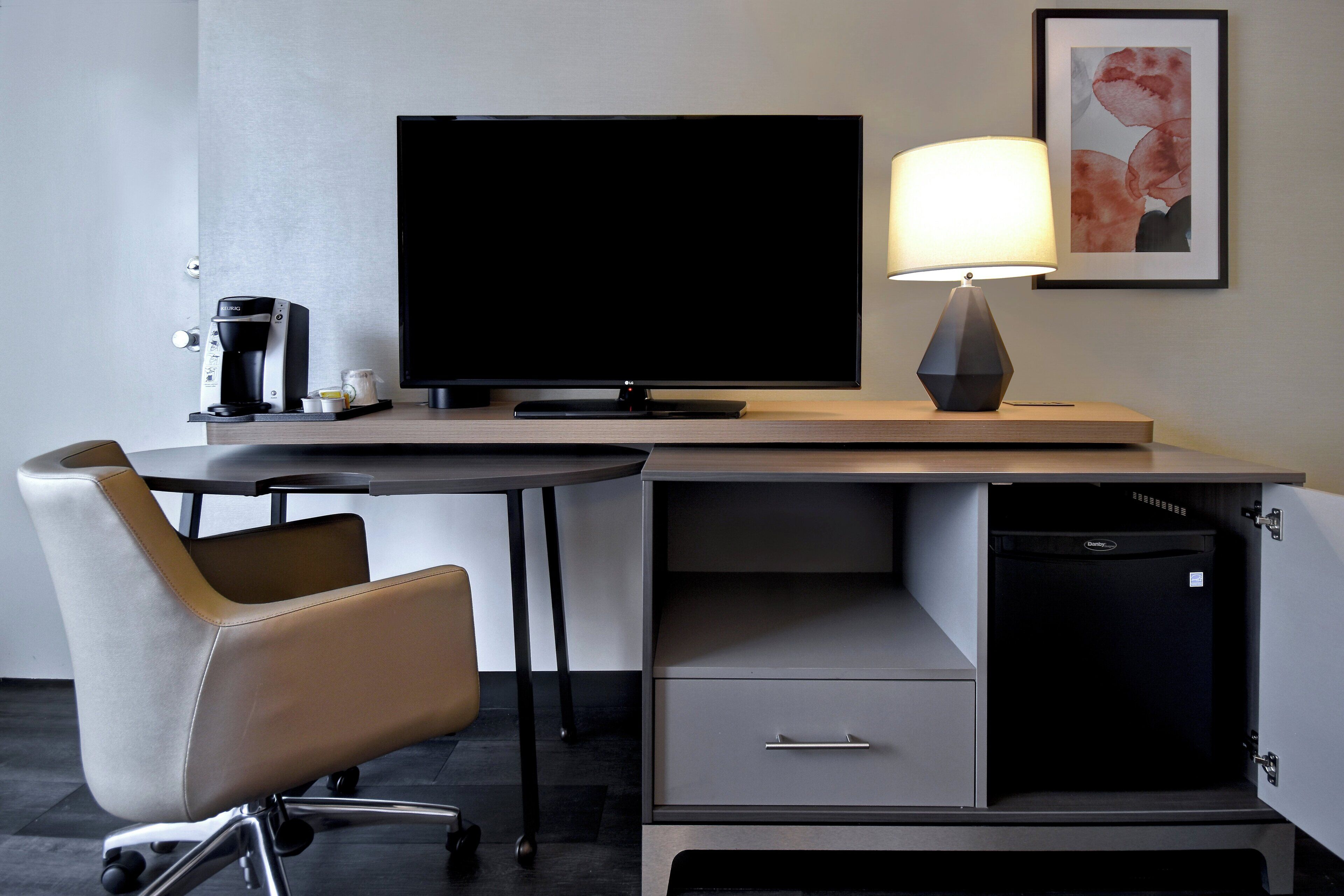 A modern hotel room with a flat-screen TV, a coffee maker, a desk with a lamp, a chair, and a small cabinet.