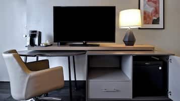 A modern hotel room with a flat-screen TV, a coffee maker, a desk with a lamp, a chair, and a small cabinet.