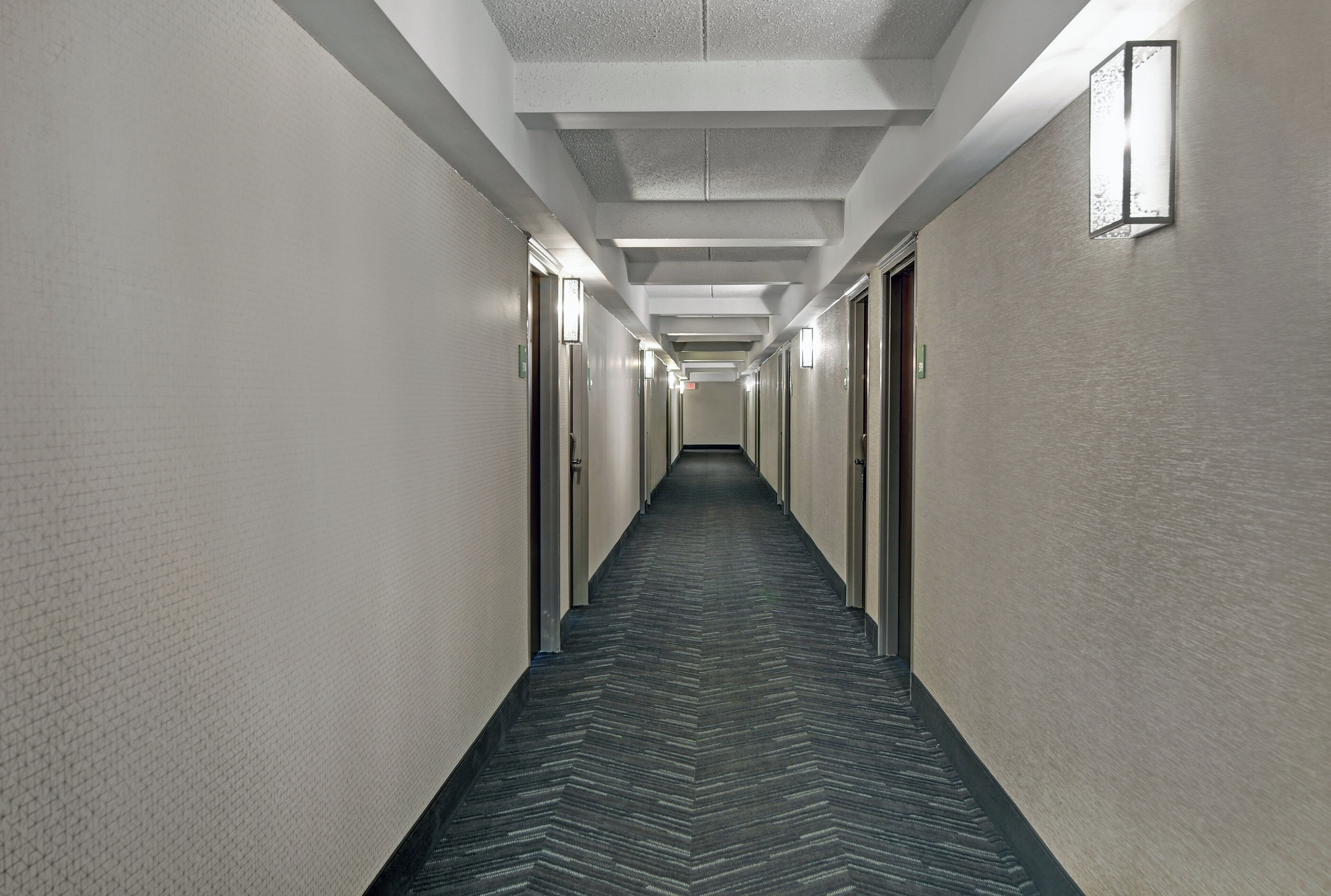 Un long couloir étroit, avec des portes de chaque côté, menant à des pièces individuelles. Le sol présente un motif décoratif et les murs sont texturés.
