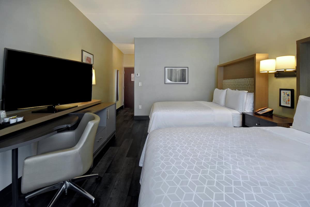 A hotel room with two beds, a desk with a flat-screen TV, a chair, and a bedside table with lamps.