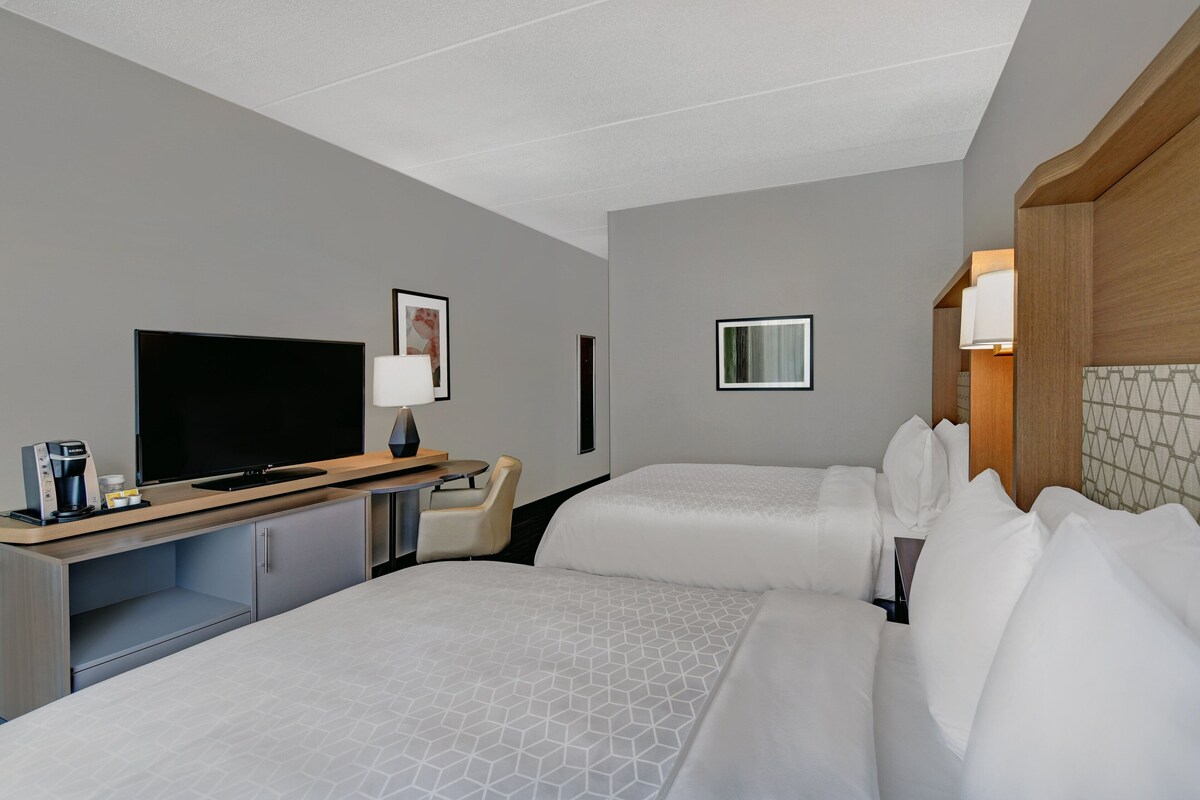 A hotel room with two beds, a TV, a desk with a coffee maker, and a chair.