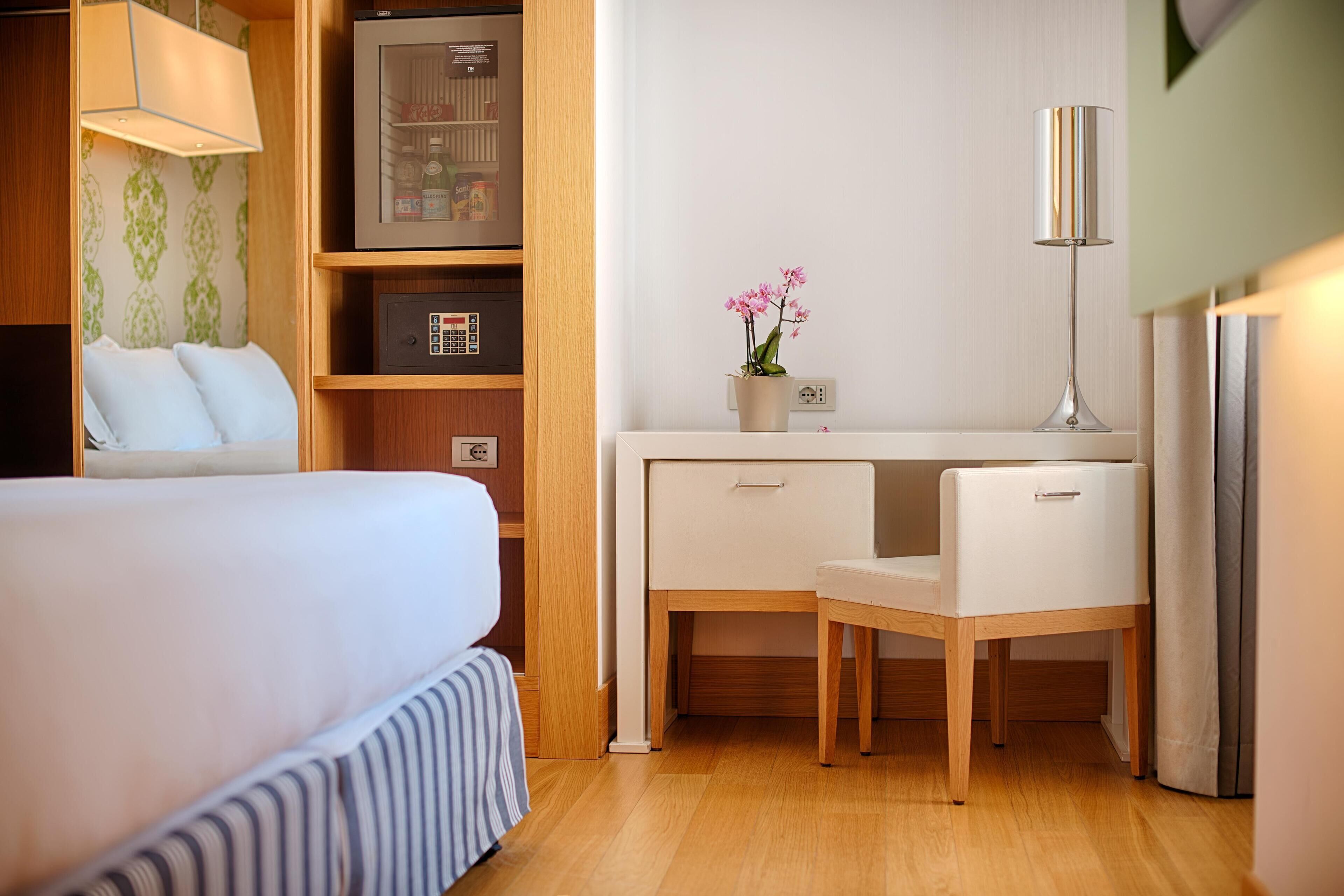 Habitación de hotel con cama, escritorio, silla, lámpara, un florero con flores y un minibar.