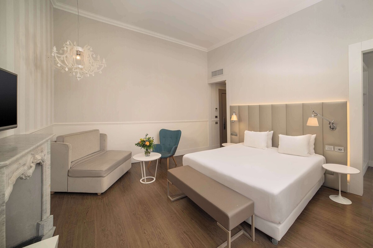 A modern hotel room with a large bed, a sofa, a small table with flowers, a bench, and a TV.