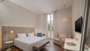 A modern hotel room with a large bed, a desk, a chair, a bench, a TV, and a chandelier. - NH Genova Centro (Genoa)