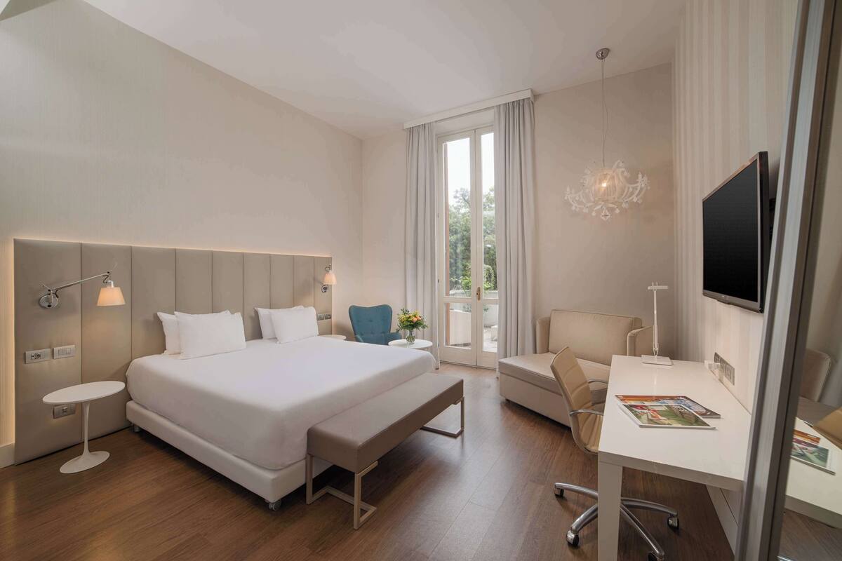 A modern hotel room with a large bed, a desk, a chair, a bench, a TV, and a chandelier.