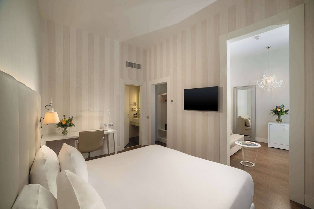 A modern hotel room with a large bed, a desk with a lamp, a TV, and a mirror.